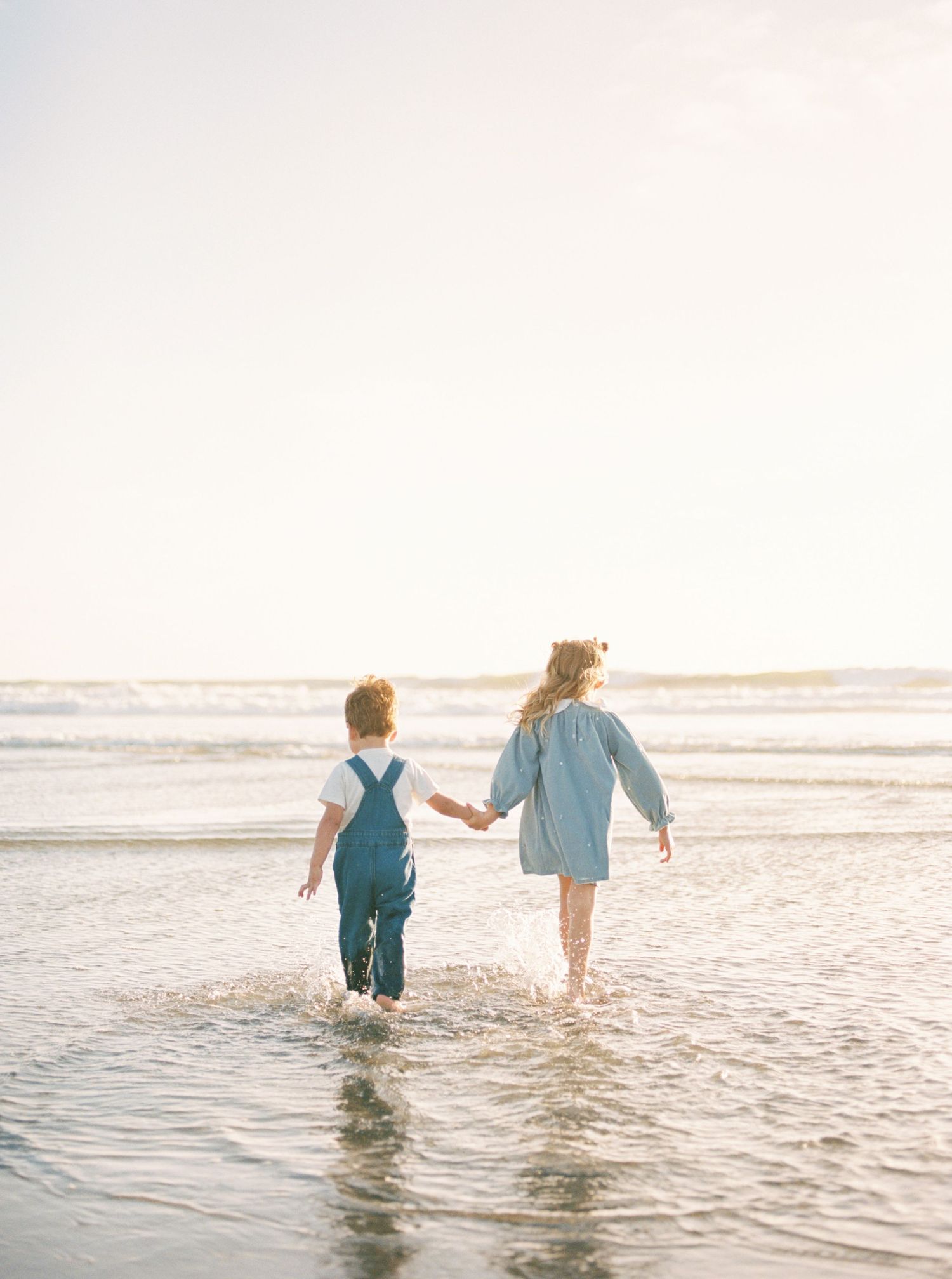 Two young children holding hands walk through shallow ocean water at the beach in soft, warm golden light.