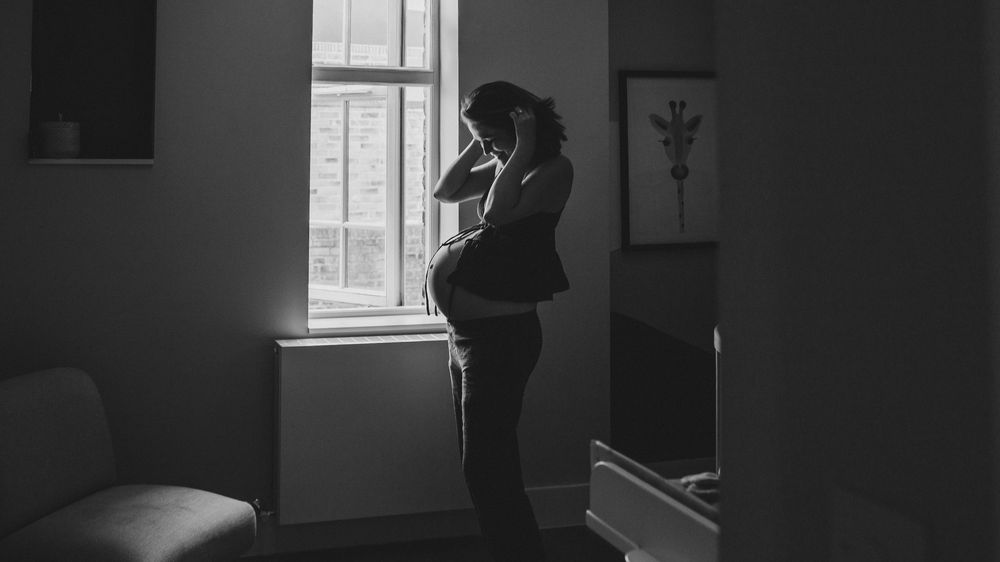 Black and white photo of a pregnant woman standing by a window