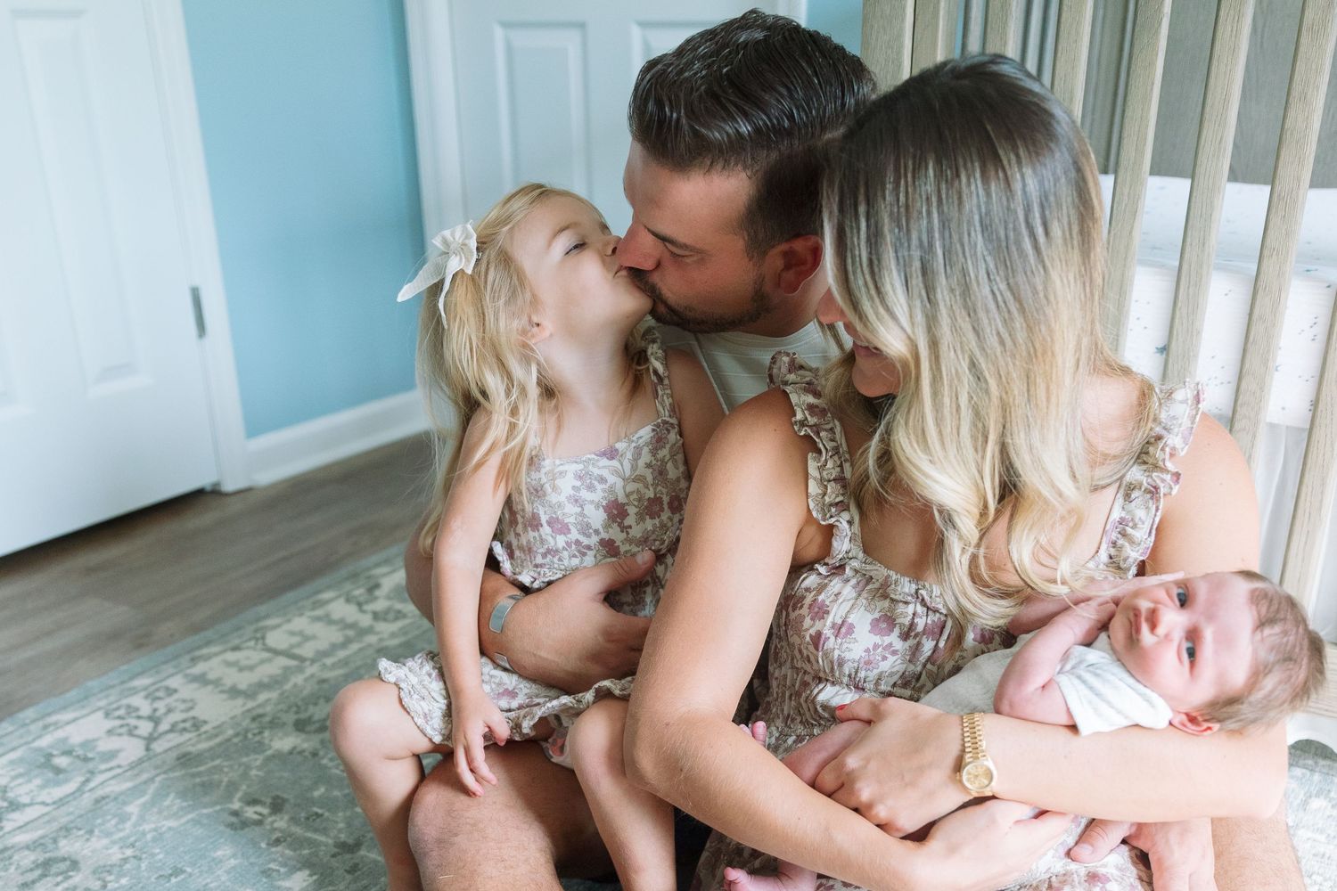 Family of four sits on floor, parents kissing while toddler girl leans in, mom holds newborn baby.
