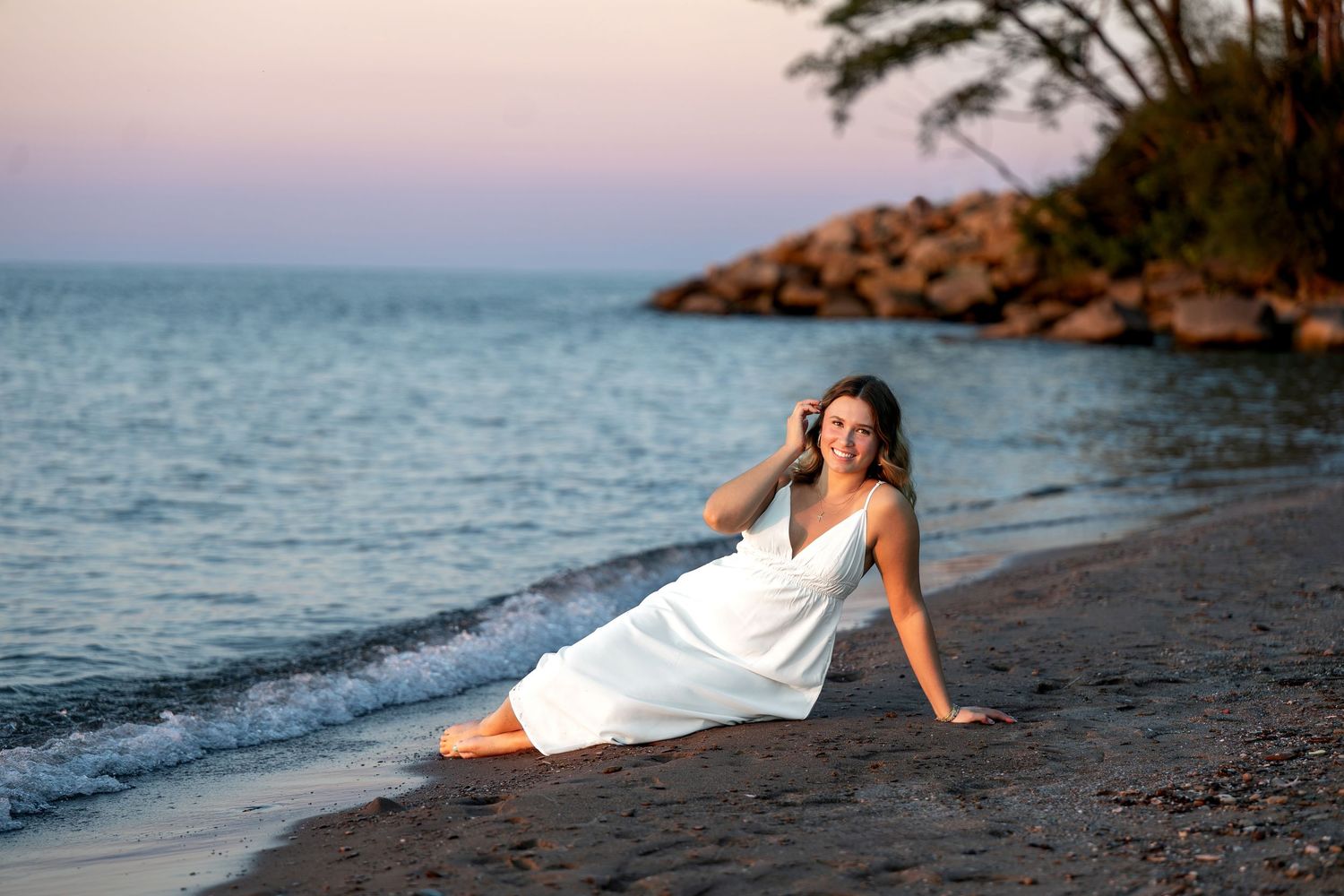 Woman in white dress sitting on sandy beach at sunset with calm ocean waves and rocky shoreline in background.