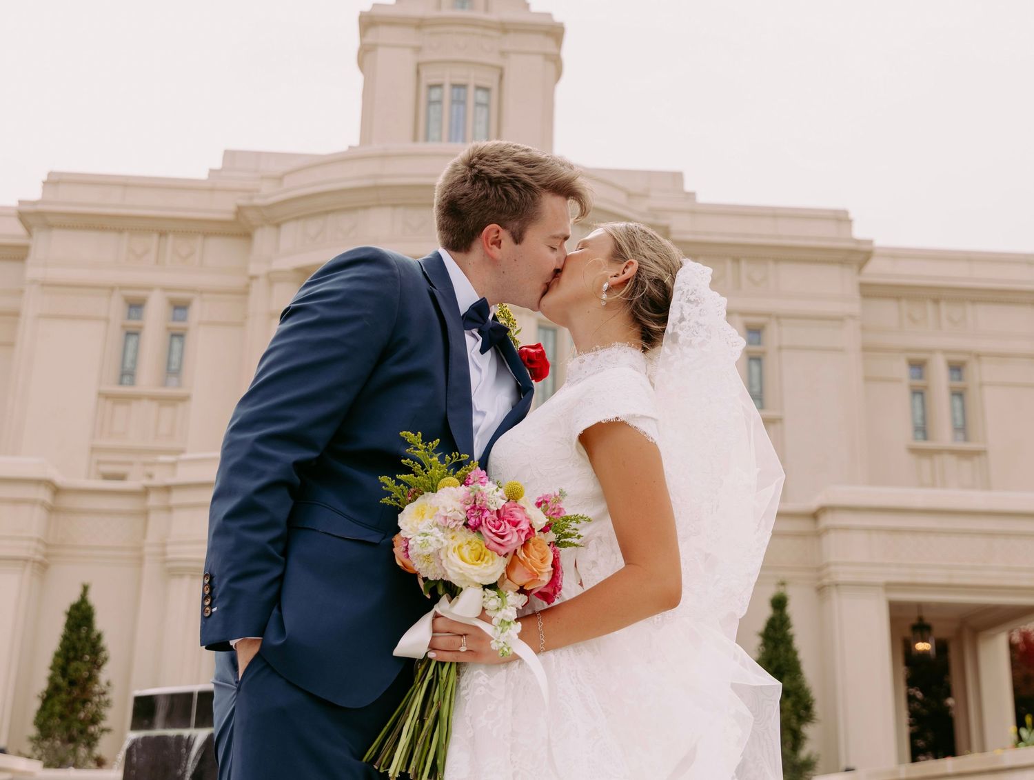 Bride and groom kissing in front of an LDS temple, bride holding colorful bouquet, groom in navy suit.