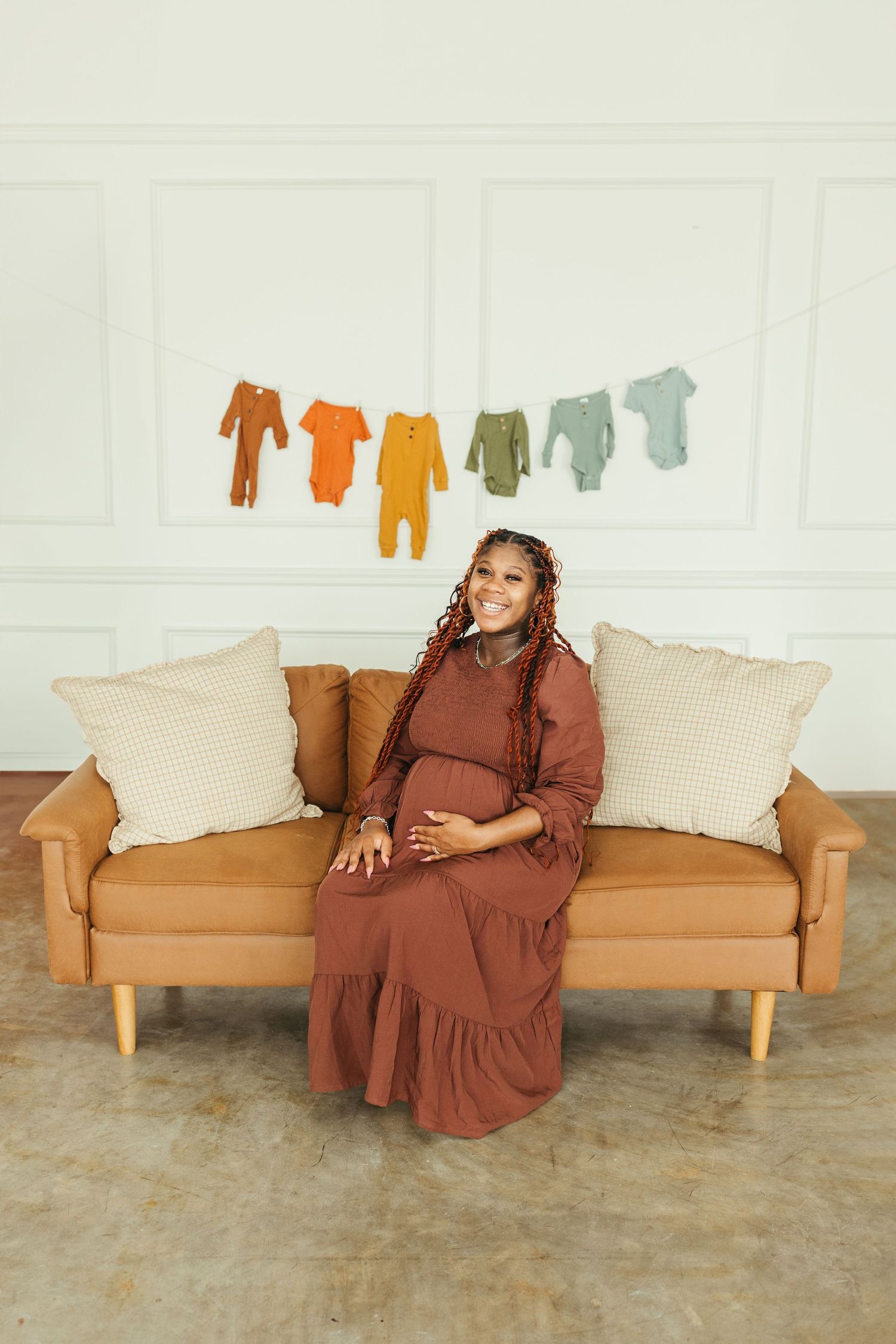 Smiling pregnant woman in brown dress sits on tan leather sofa with baby clothes hanging on string behind her.