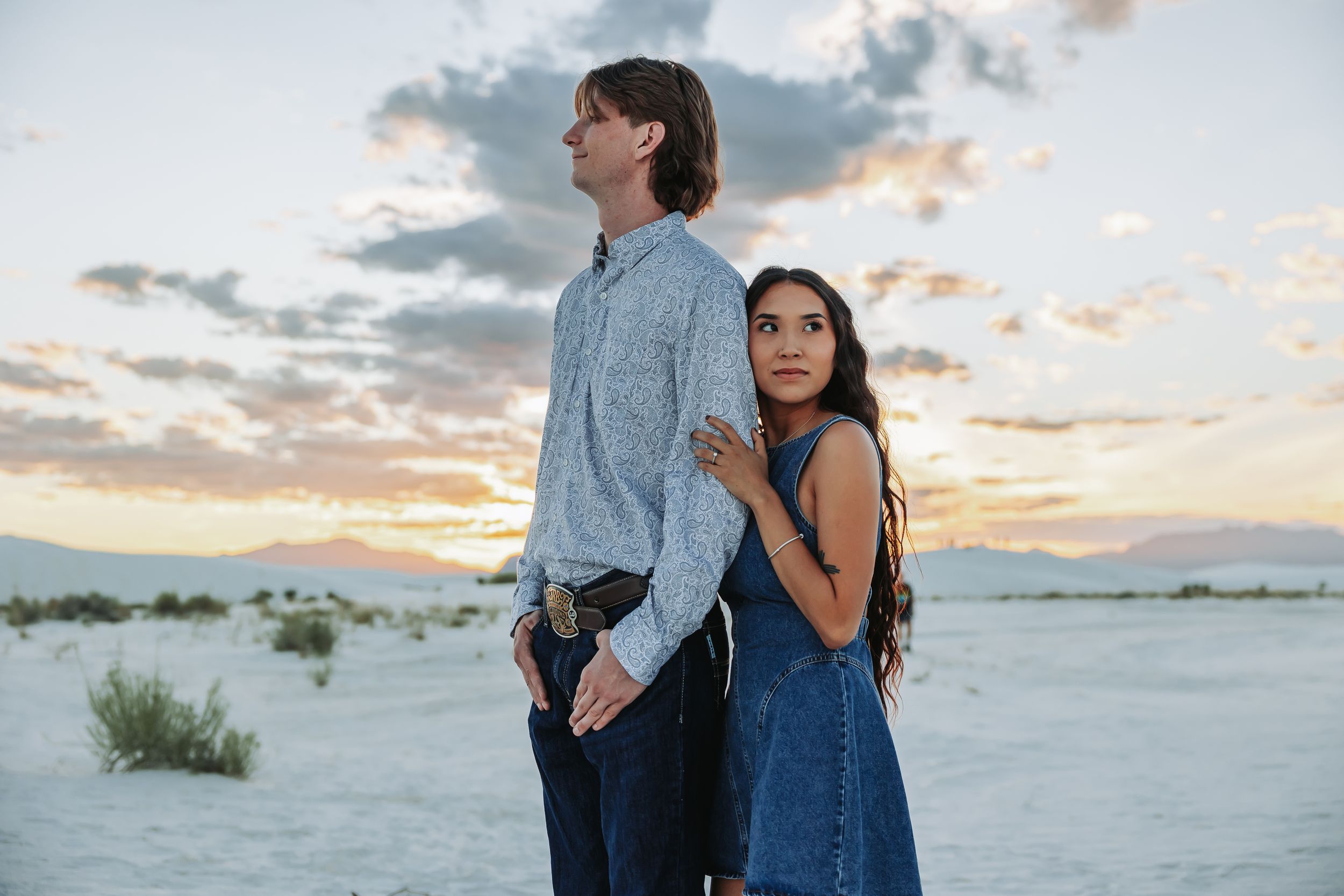 Couple standing back-to-back in white sand desert at sunset, woman in blue dress, man in gray shirt.