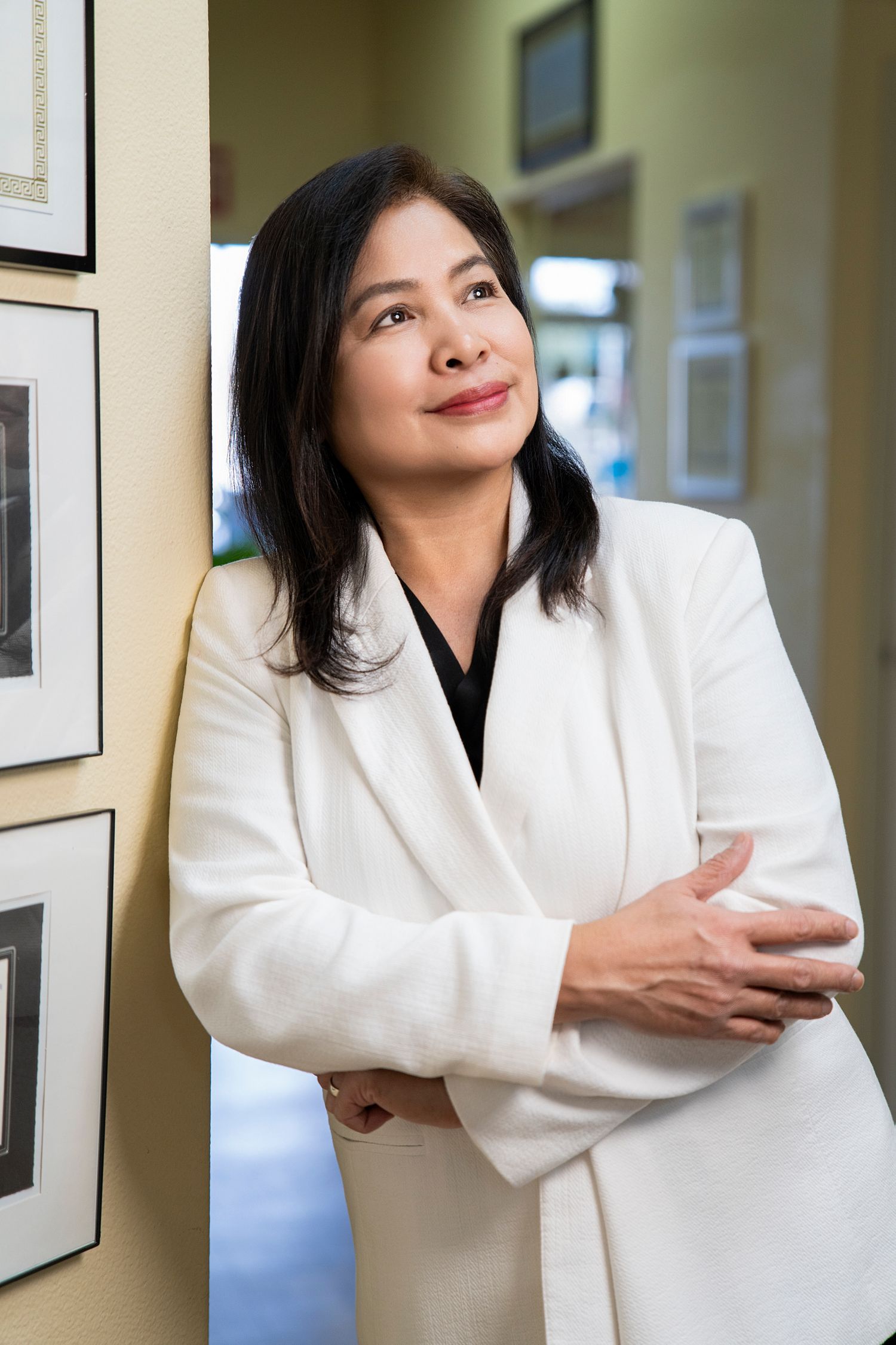 Professional Asian woman in white blazer posing confidently in an office hallway with framed pictures on the wall.