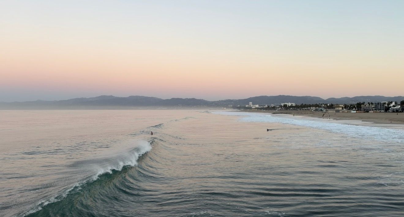 Gentle ocean waves roll toward a sandy beach at sunset, with a surfer in the water and distant mountains under a soft pink and blue sky.