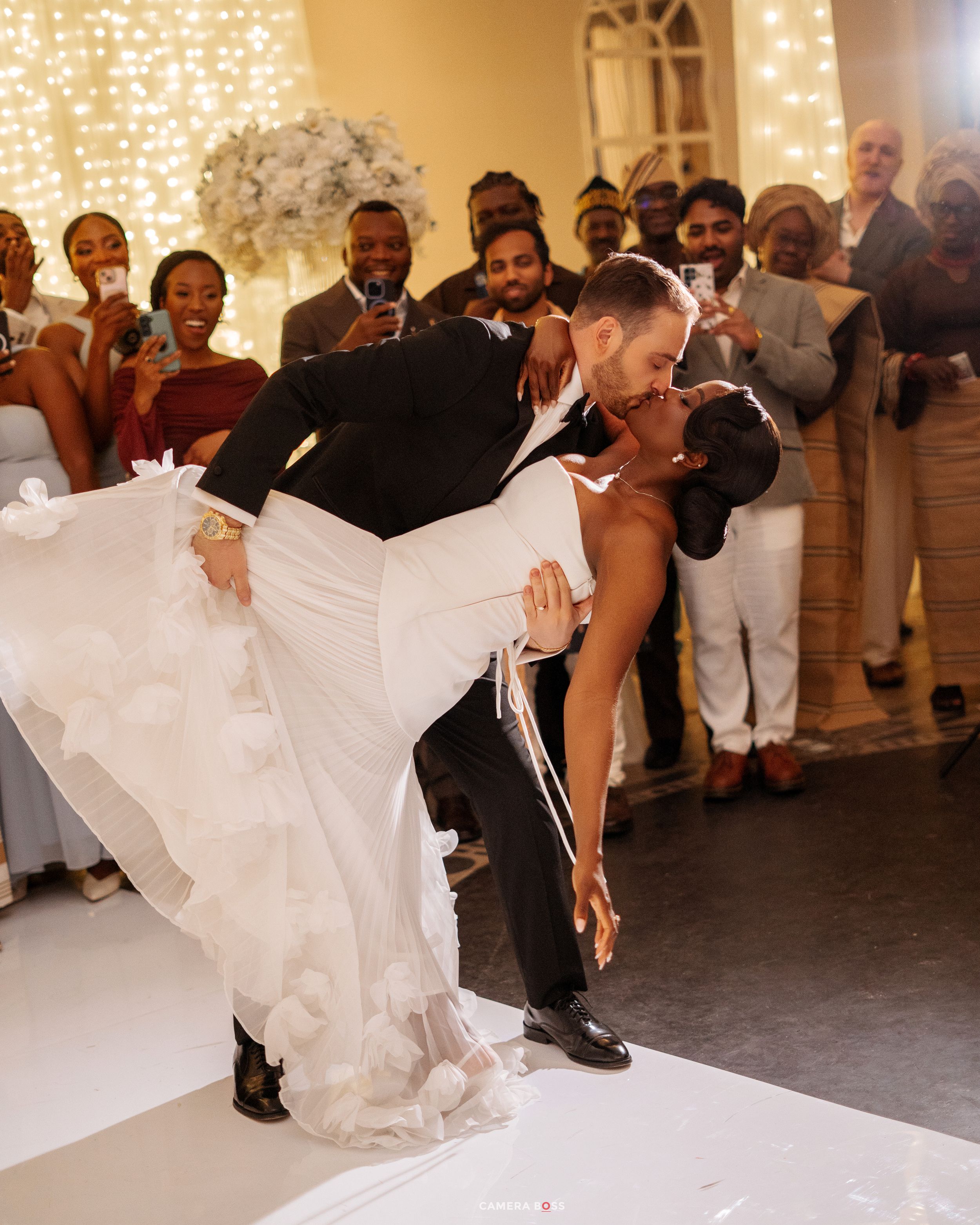 Groom dips bride in elegant white wedding gown during romantic first dance, surrounded by cheering guests at reception.