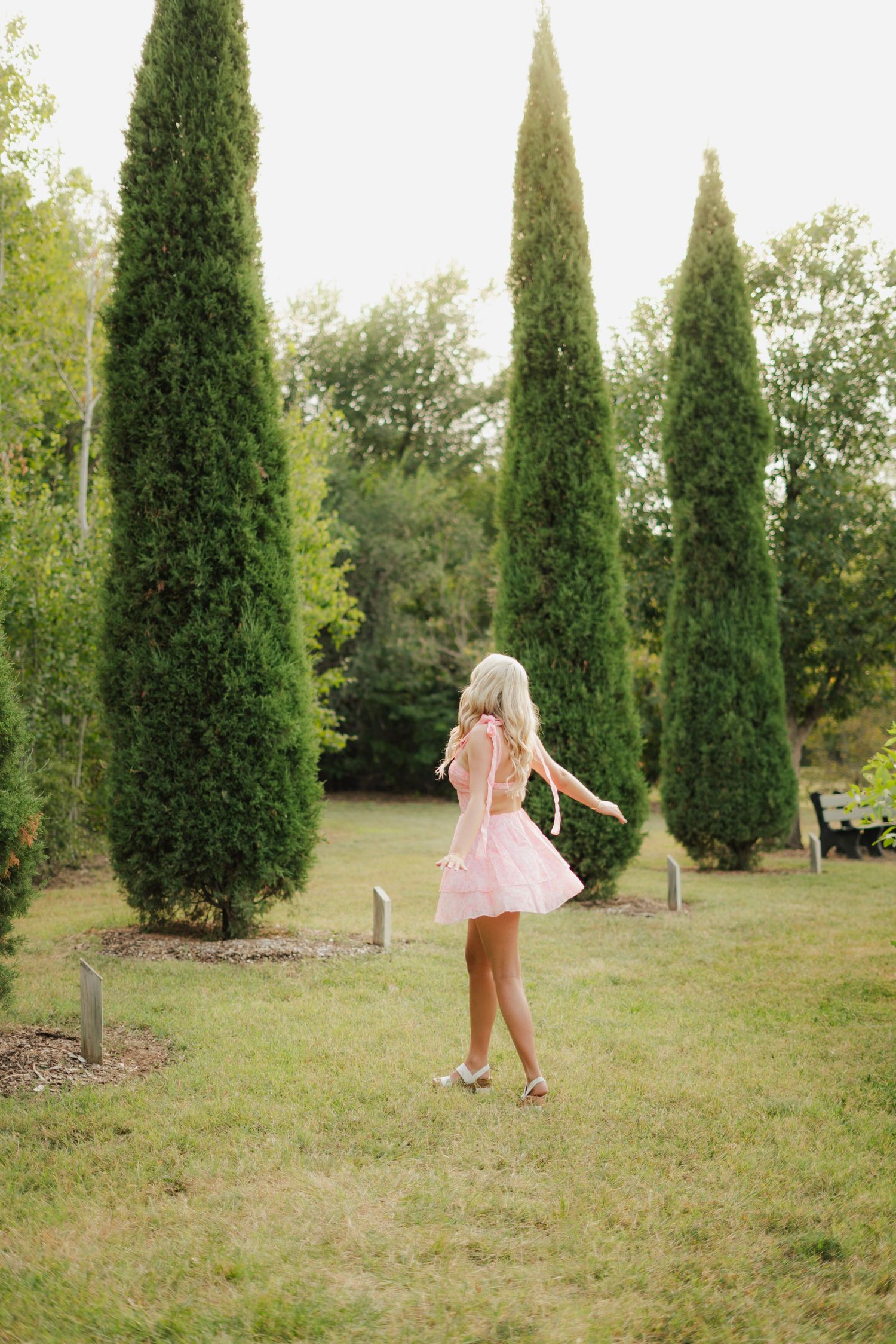 Blonde woman in mint dress sitting on wooden dock by calm lake surrounded by lush green trees.