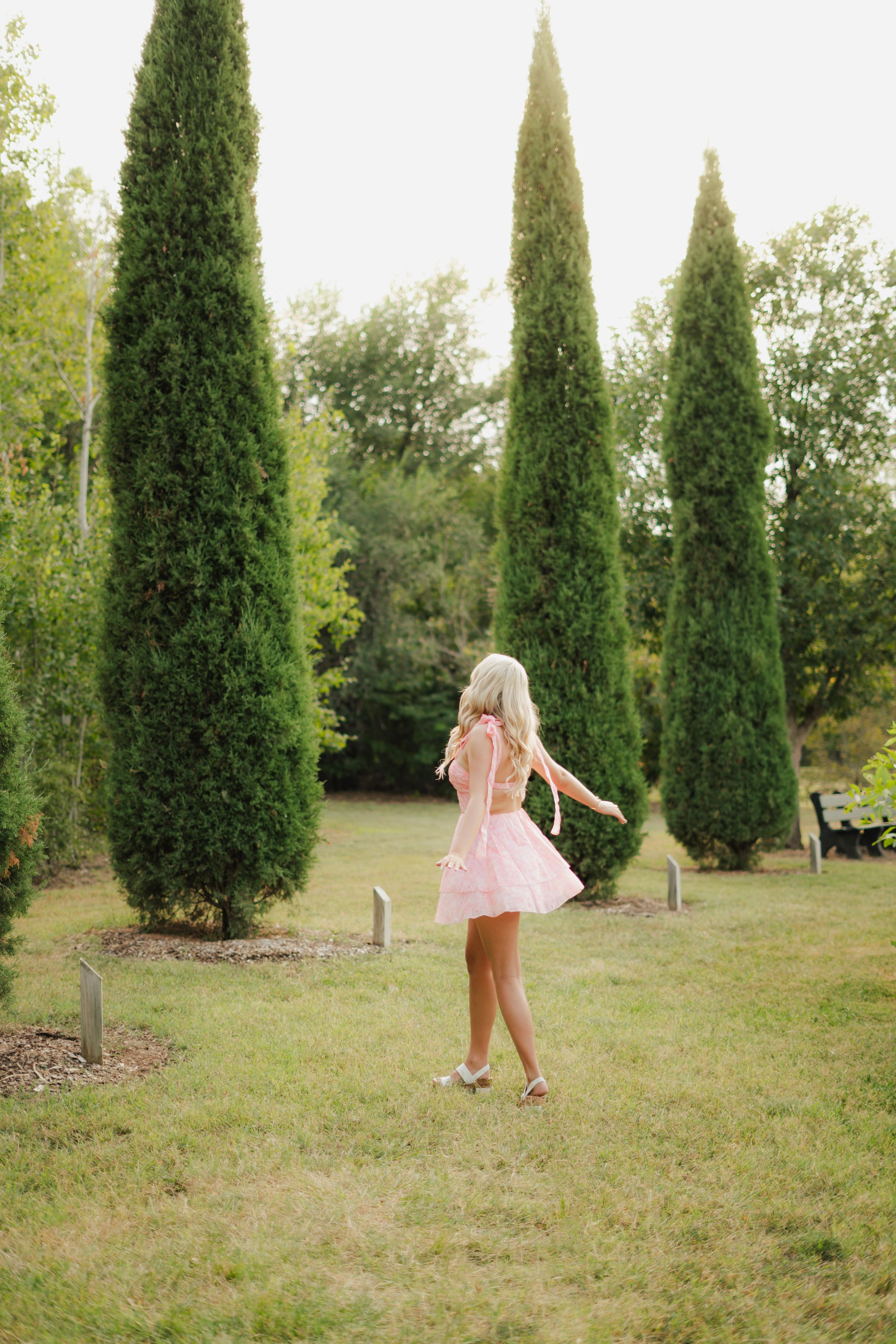 Blonde woman in mint dress sitting on wooden dock by calm lake surrounded by lush green trees.
