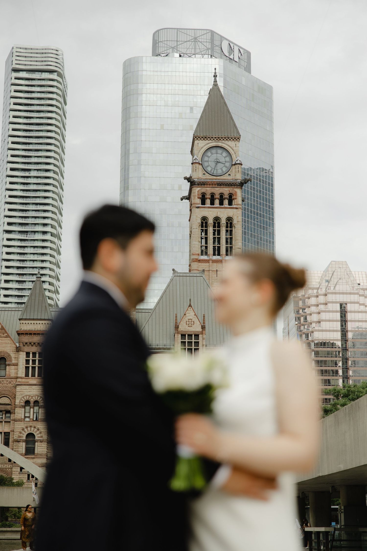 Bride and groom stand in front of Toronto City Hall clock tower and modern skyscrapers on their wedding day.