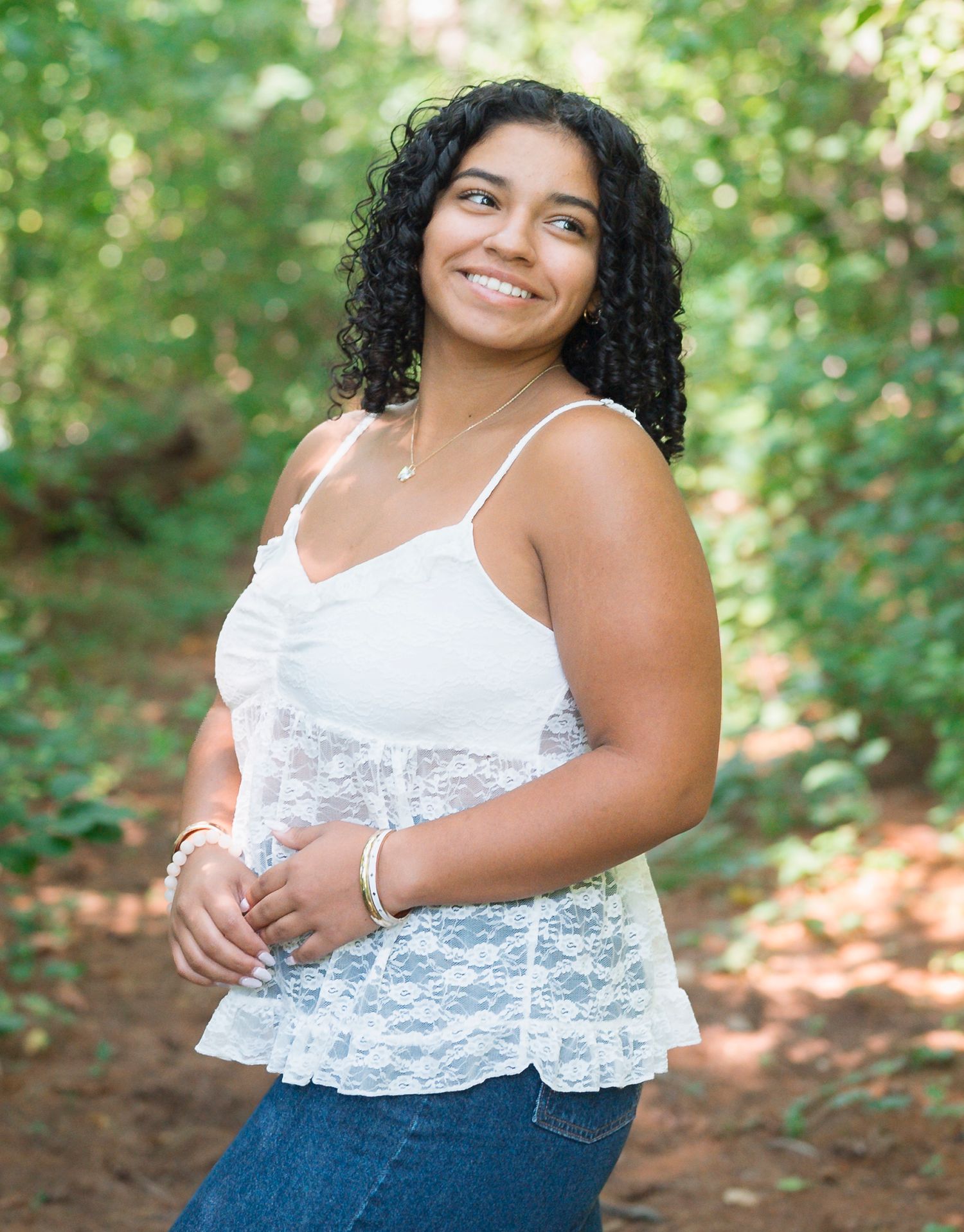 Young woman with curly hair smiling outdoors, wearing a white lace top and jeans in a lush green forest setting.