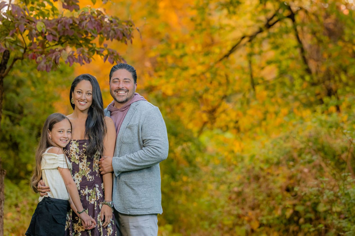 Smiling family of three posing together outdoors surrounded by vibrant orange and yellow autumn foliage in a sunny park.