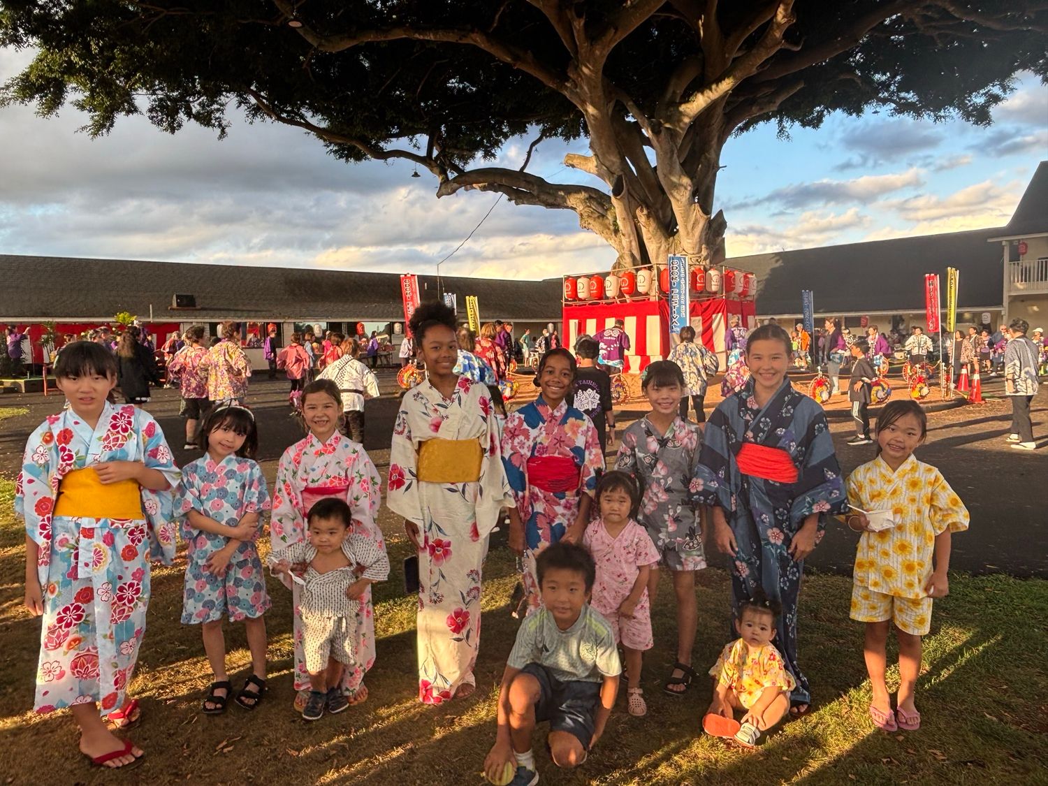 Children in colorful yukatas pose together outdoors at a Japanese festival with red lanterns and a large tree.