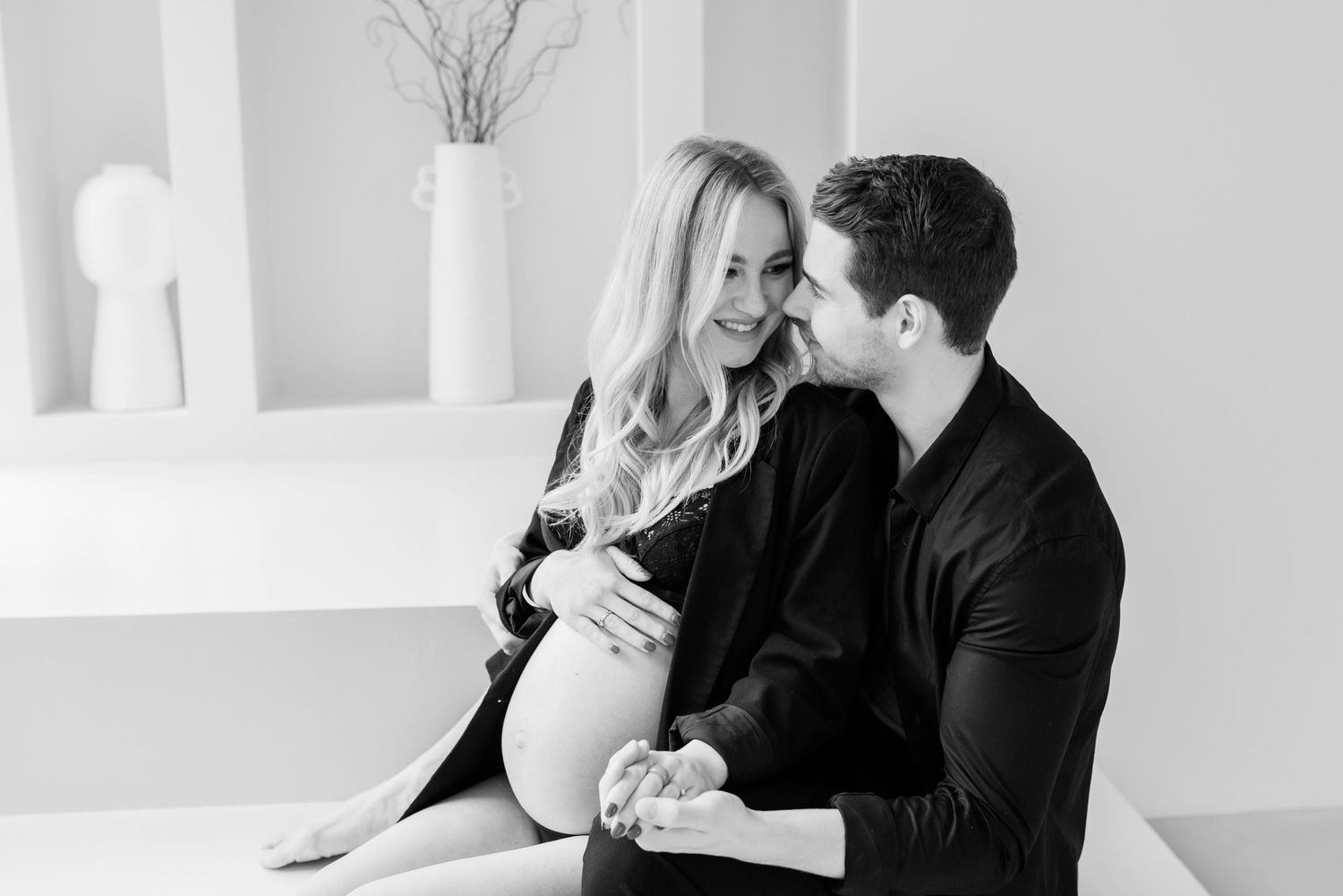 Romantic couple shares an intimate moment in black and white while sitting together in a minimalist white room.