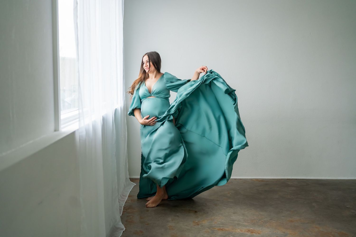 A maternity portrait with a woman in a flowing teal dress dancing in a bright studio setting.