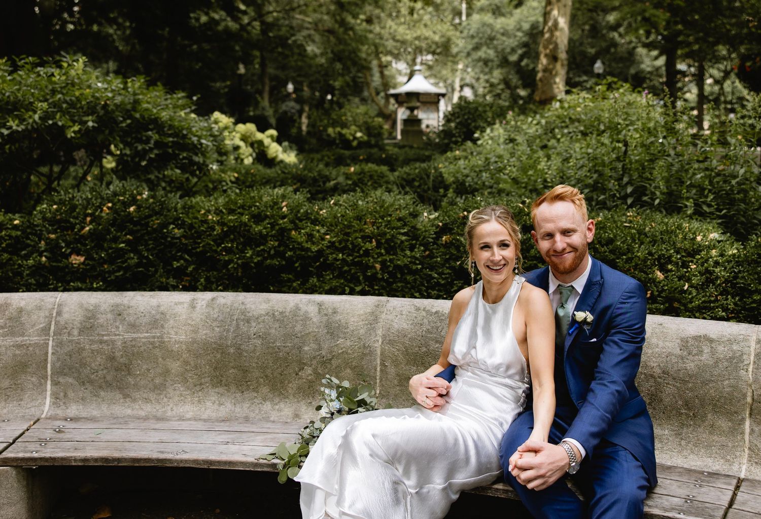Wedding couple posing on a stone bench surrounded by lush greenery. Rittenhouse Square Philadelphia, PA.