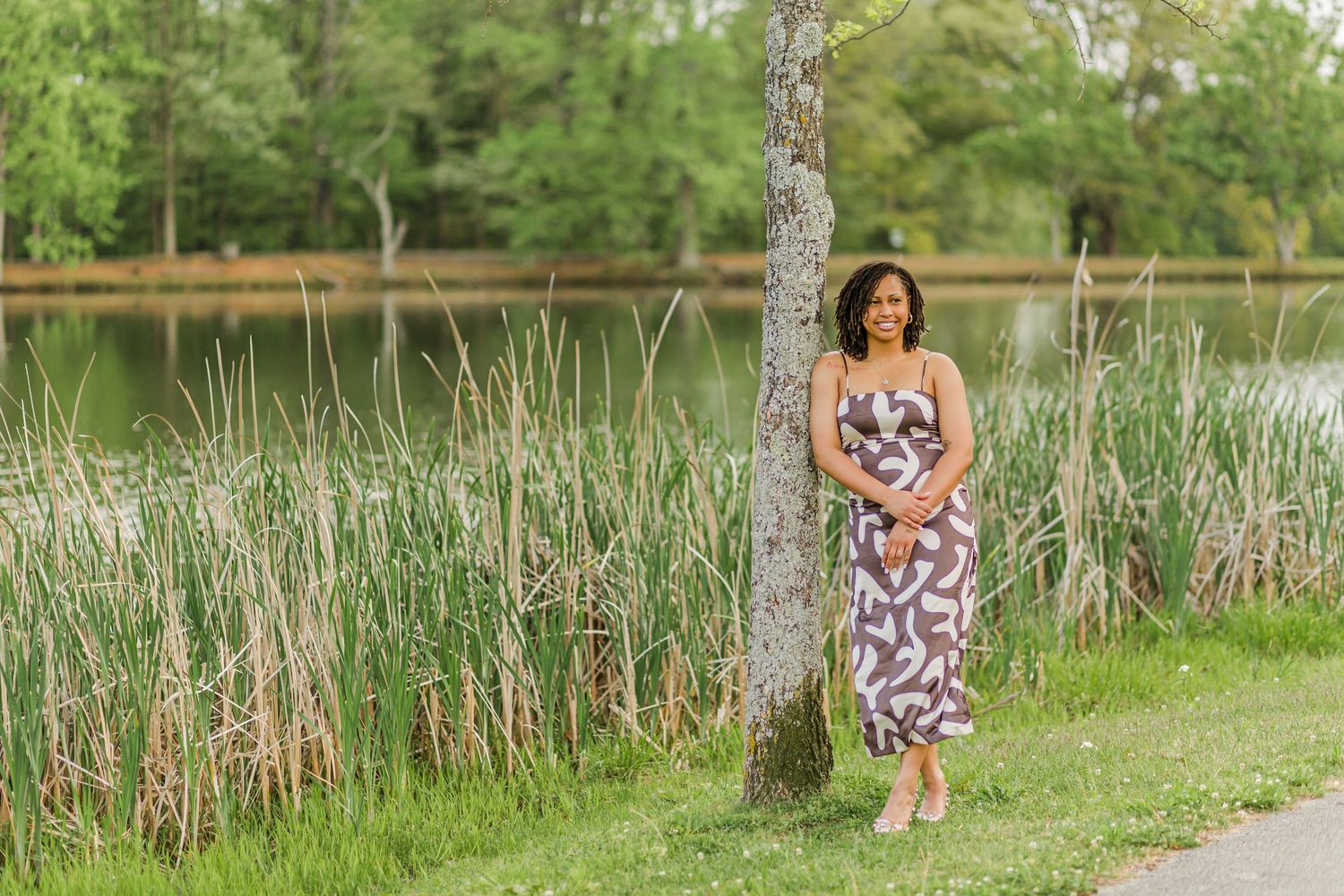 Woman in floral dress poses by lake with cattails and trees in background.