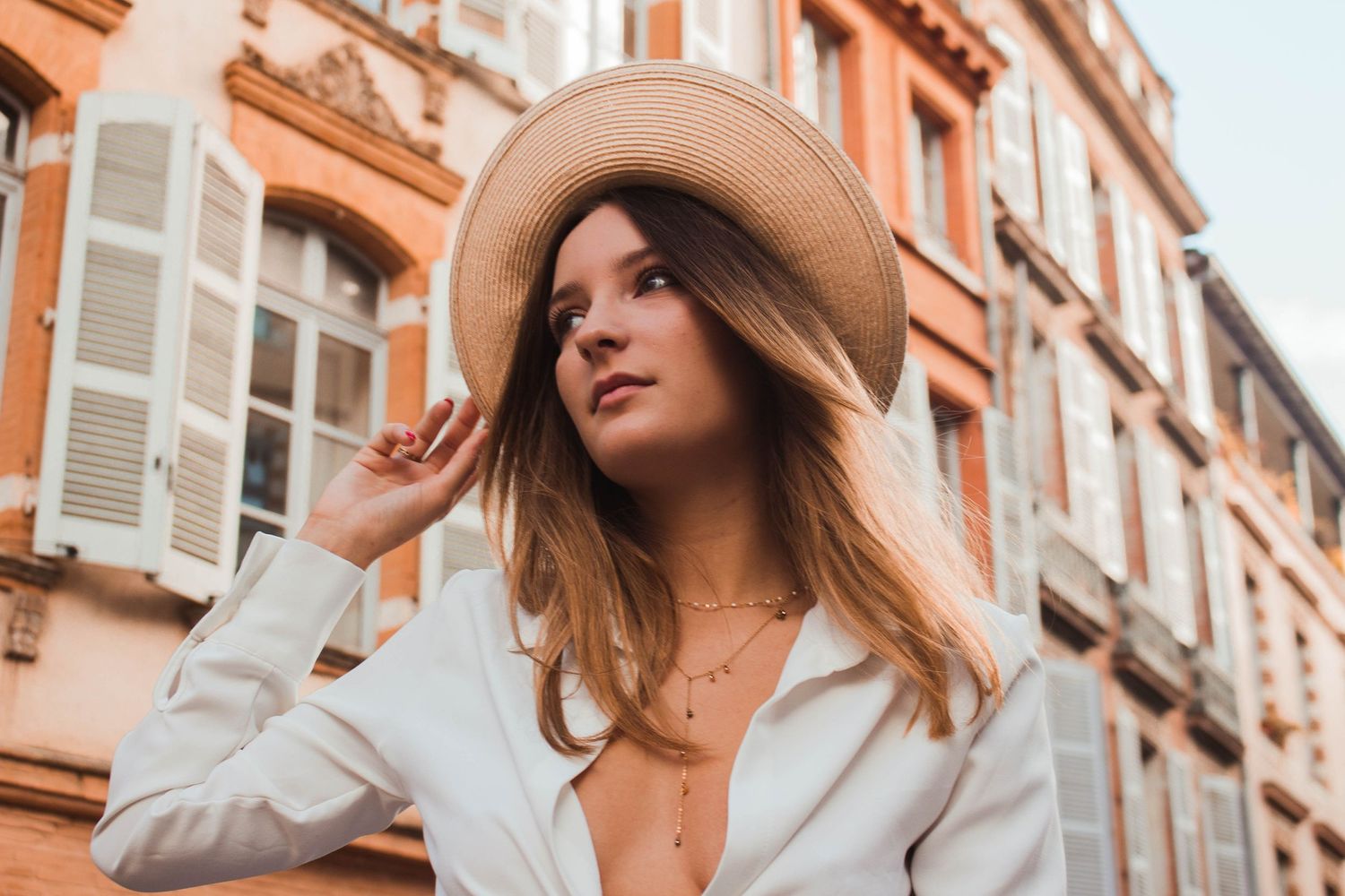 Stylish portrait of someone in a white blazer and straw hat against orange buildings in European city.