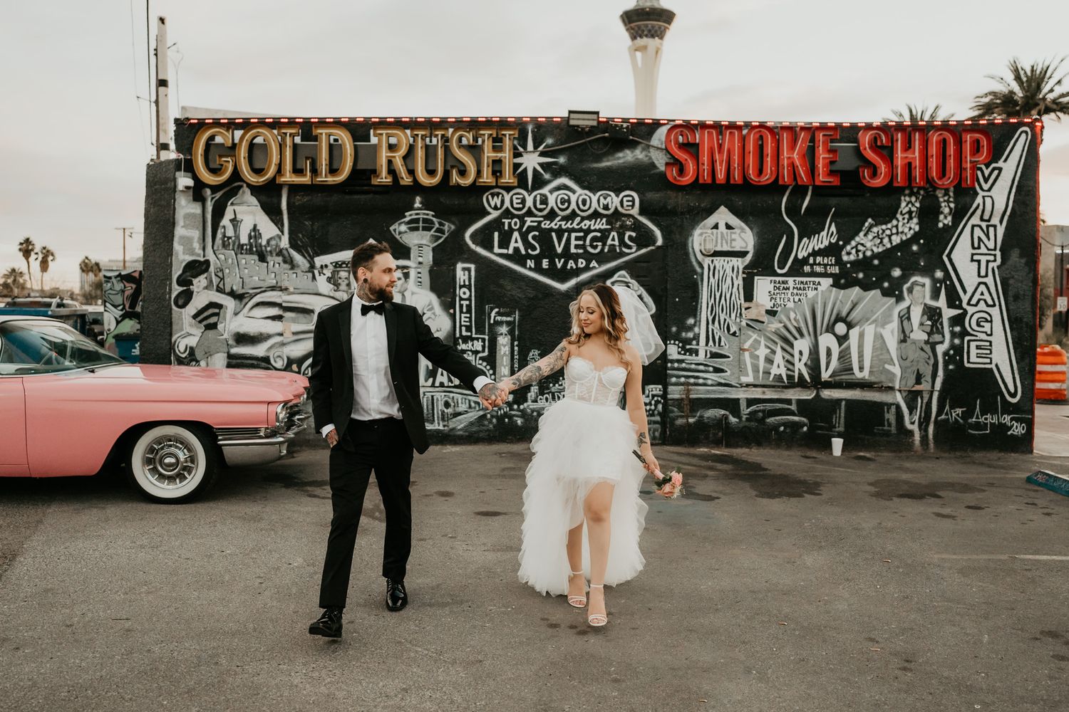Newlyweds pose in front of retro Gold Rush smoke shop mural with vintage pink car.