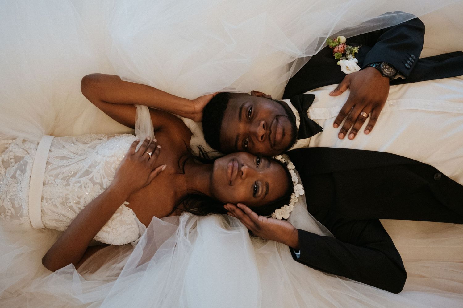 A wedding couple in formal attire lies together on white fabric sharing a joyful moment.