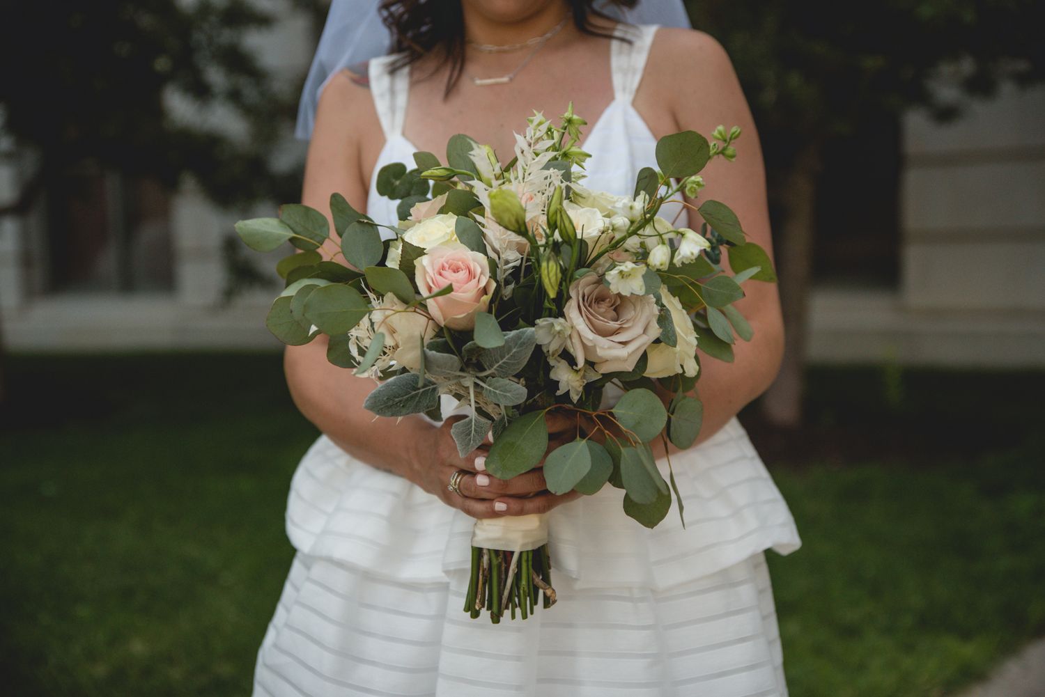 A bridal bouquet featuring delicate blush roses and greenery held against a white layered dress.