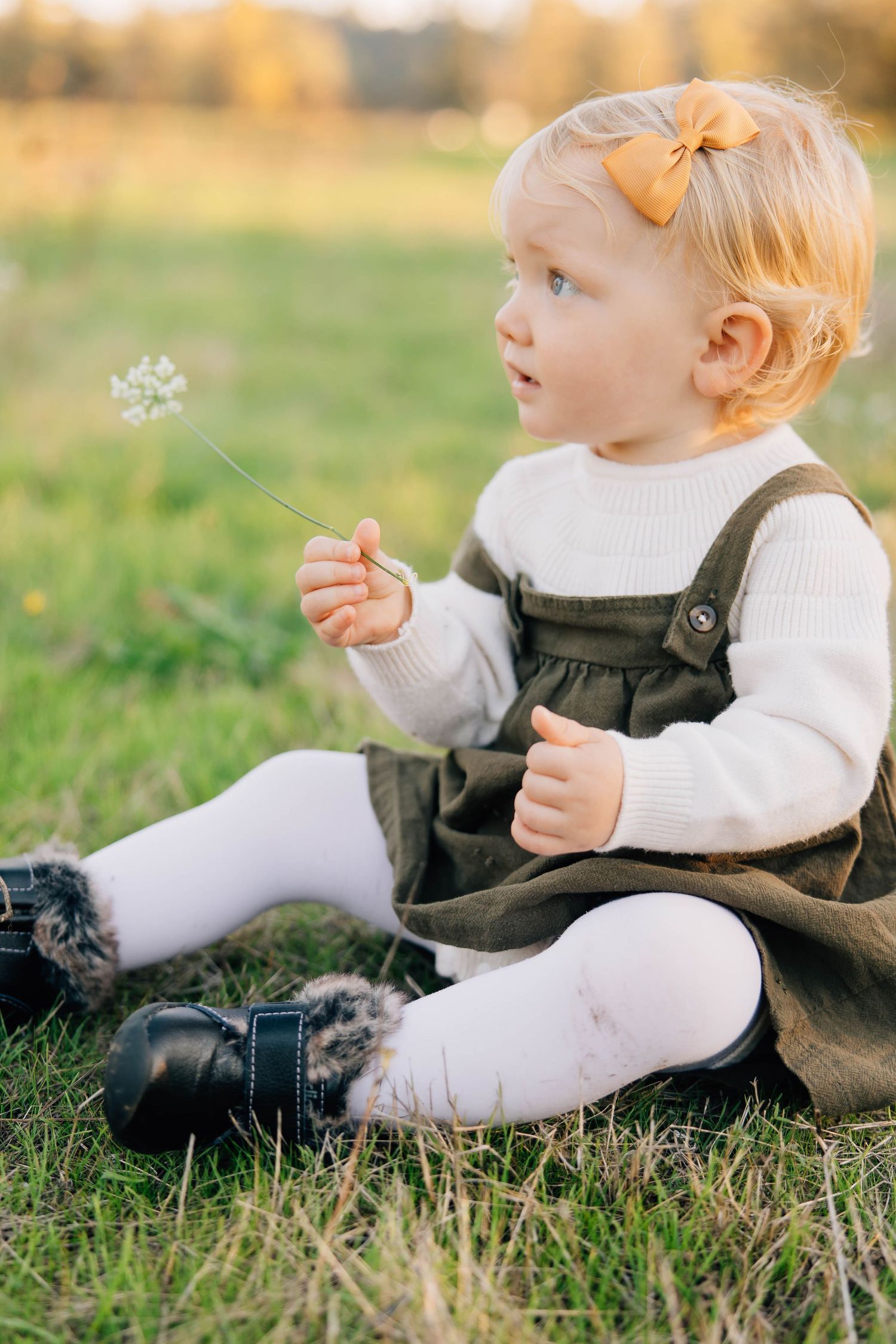 Adorable blonde toddler girl in olive dress sitting on grass holding a wildflower.