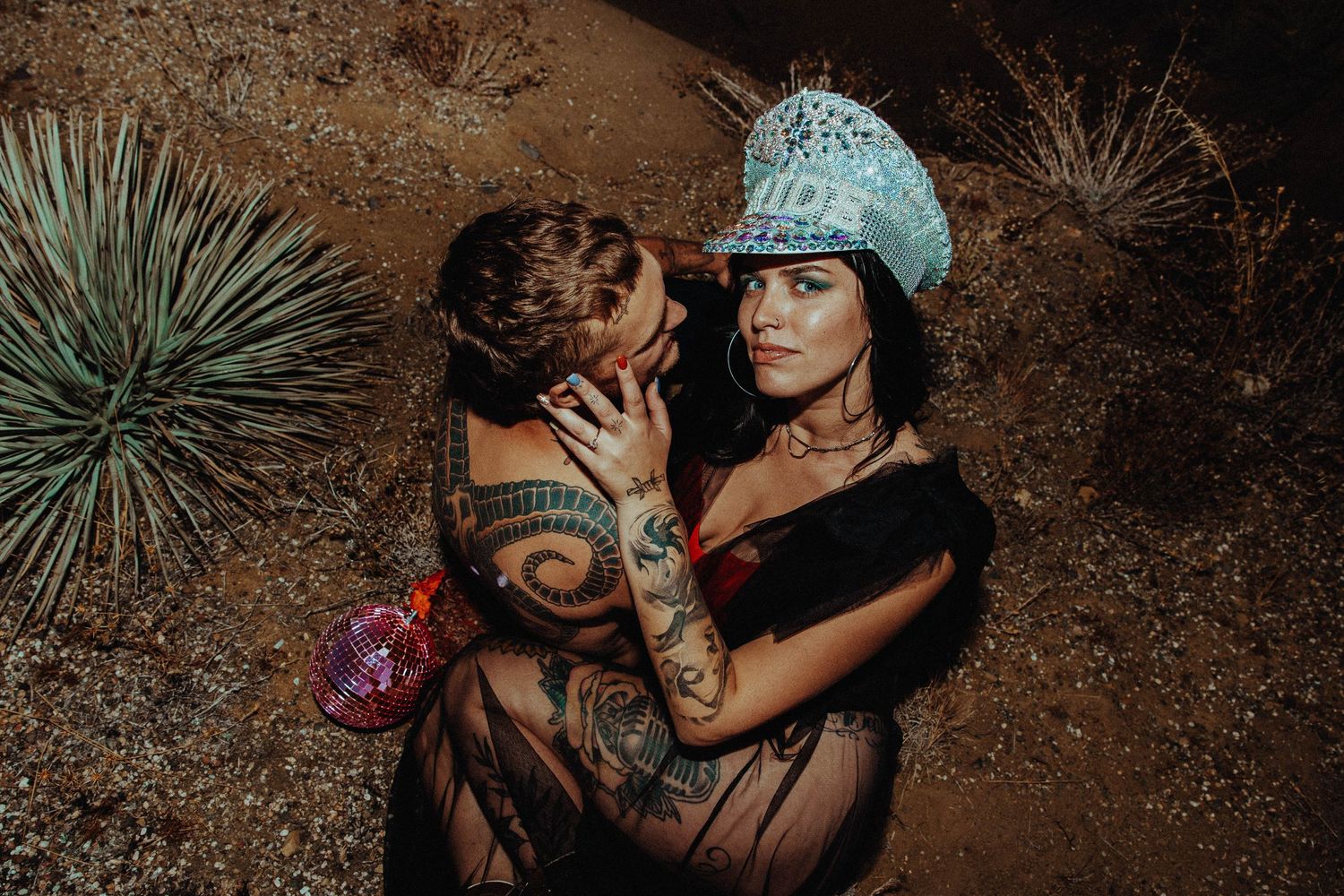 A stylistic portrait of a couple embracing in the desert with yucca plants and tattoos visible in moody lighting.