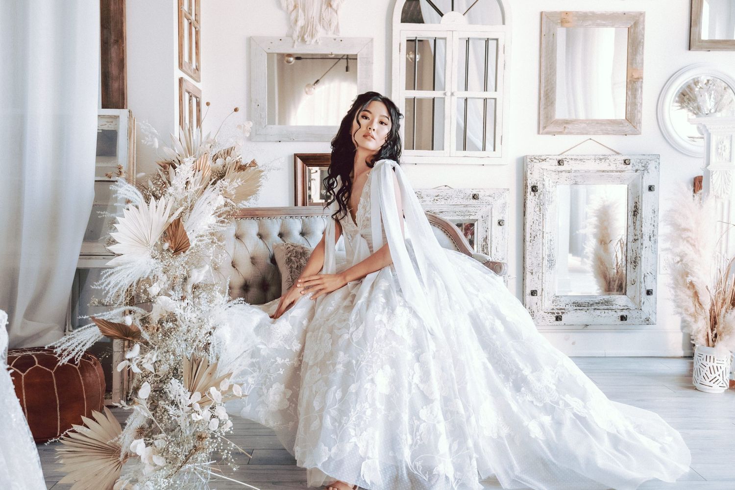 A bride in a flowing white floral gown poses gracefully in a boho-styled room with dried pampas grass.