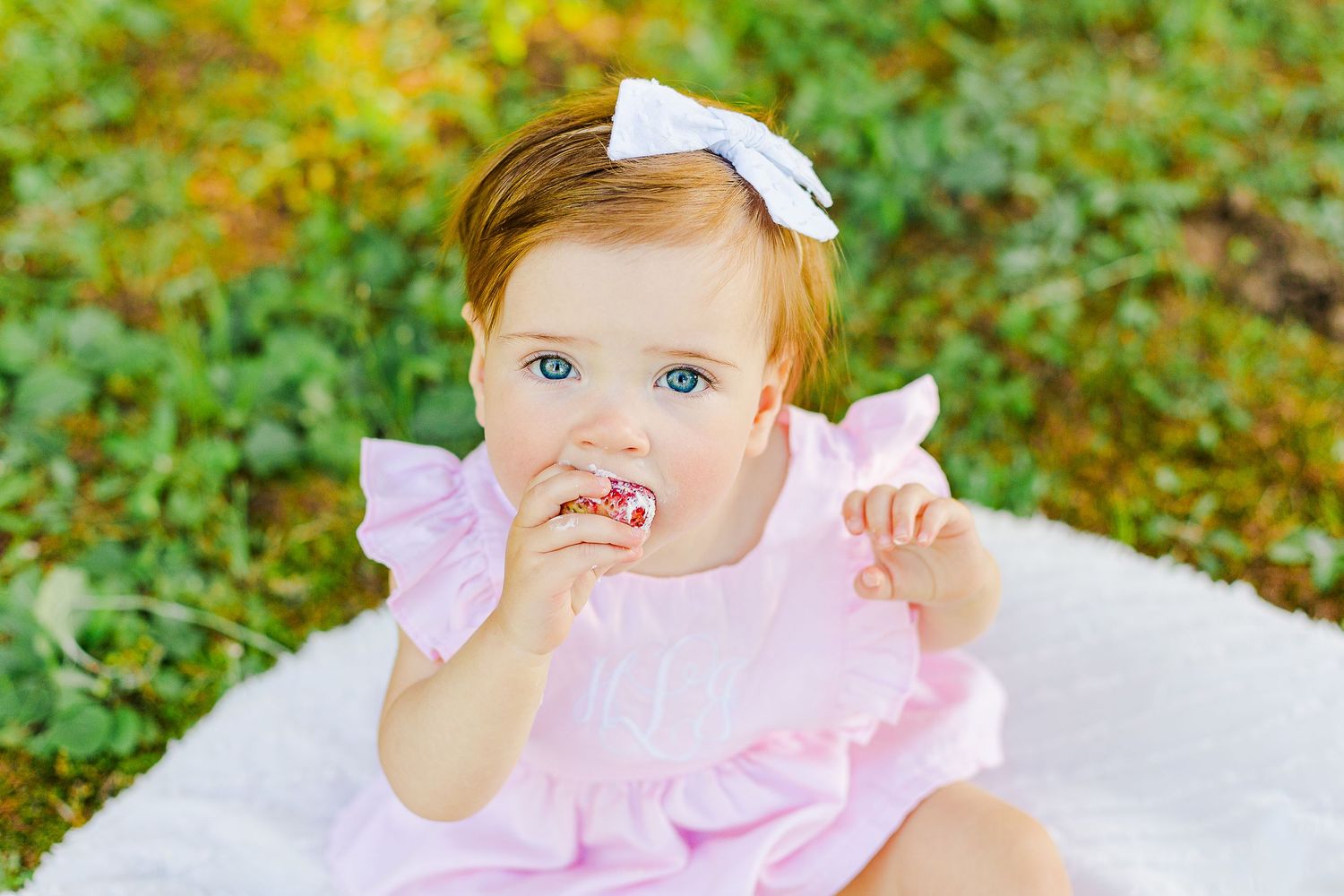 A small child in pink wearing a white bow sits on a blanket outdoors.