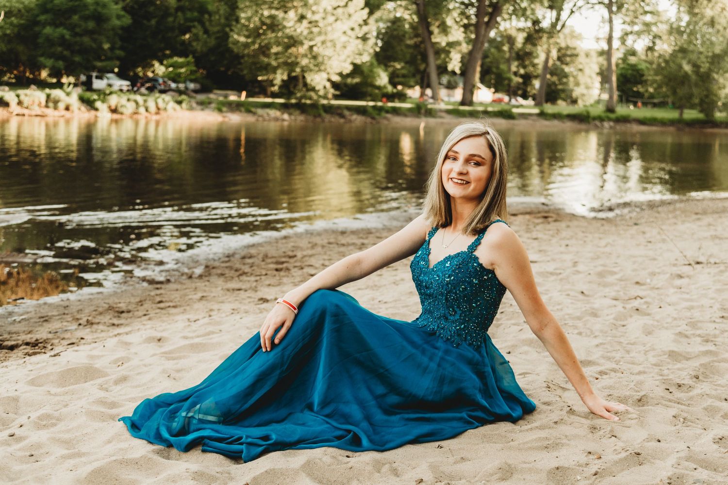 Woman in elegant teal evening gown poses on sandy beach near water.
