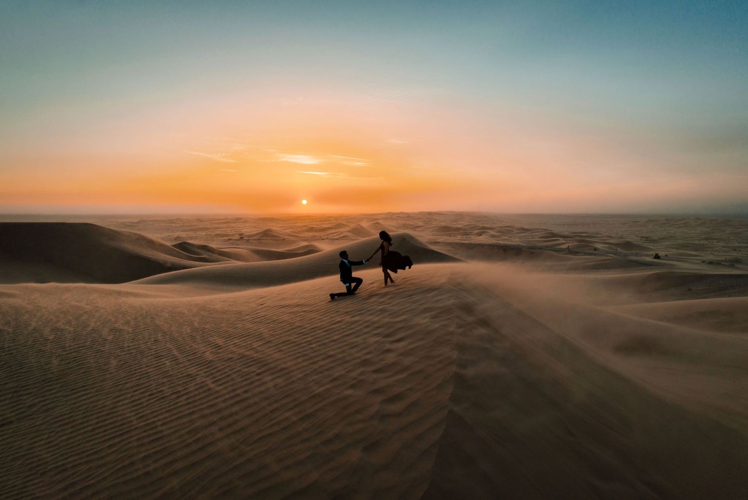 Two silhouettes walking across sand dunes during a golden desert sunset.