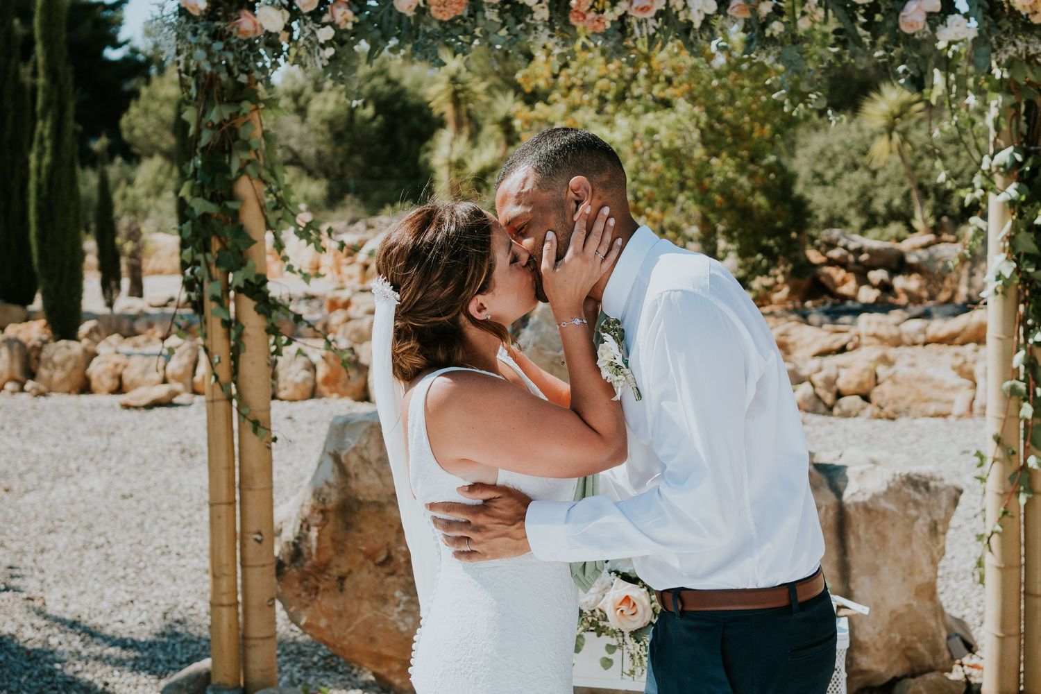 A tender moment between newlyweds under a floral arch in a rustic outdoor setting.