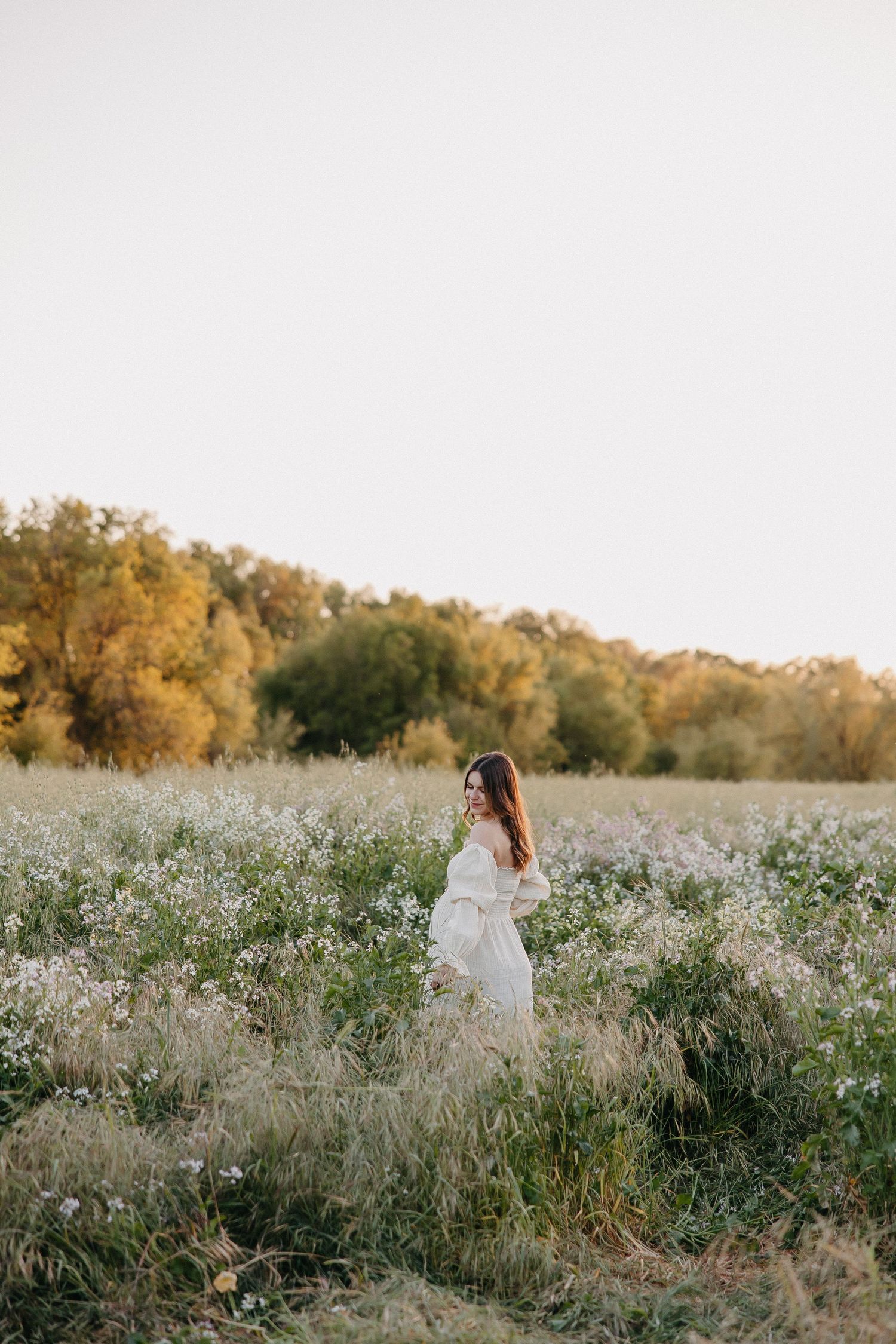 A person in a flowing white dress walks through a field of wildflowers with autumn trees in background.