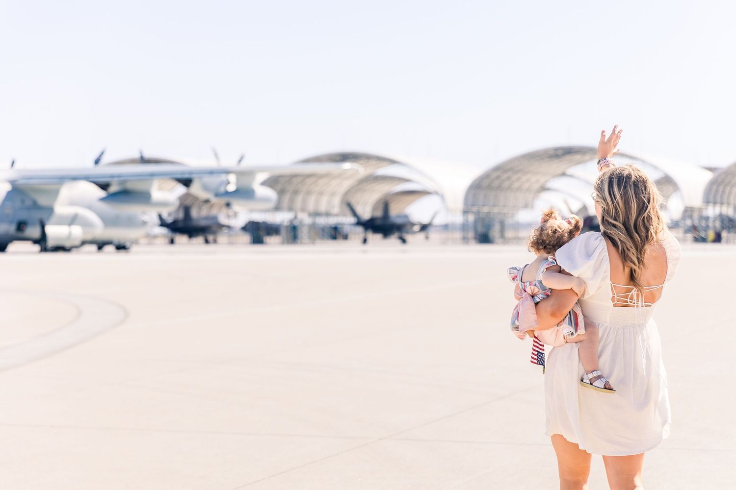 Woman in white dress carrying child walks across airfield with aircraft hangers in background during sunset.