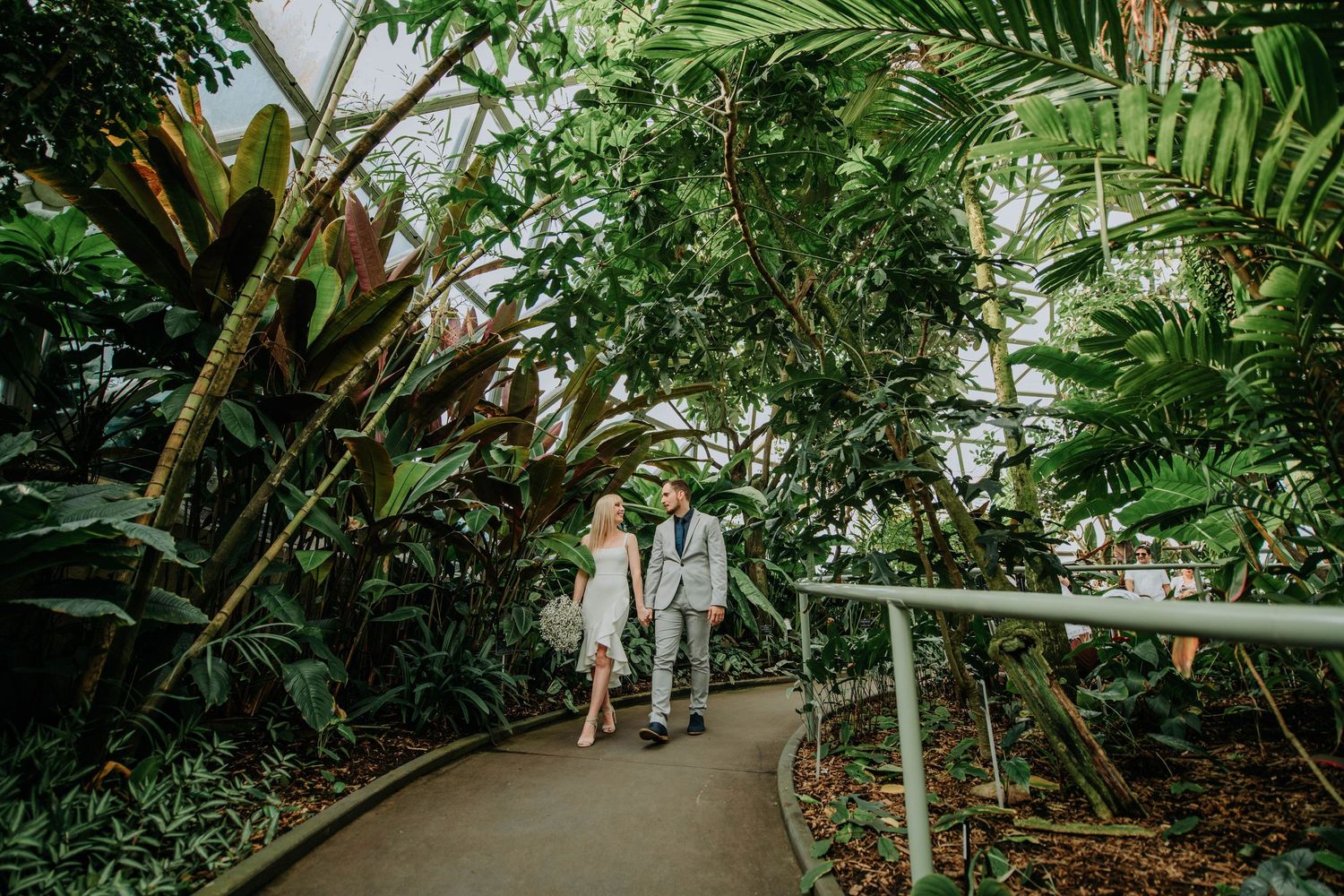 Couple walking along a curved pathway in a lush tropical greenhouse or conservatory.