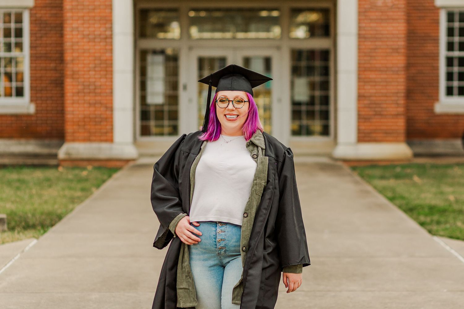 Graduate in cap and gown poses in front of brick building with bright pink hair.