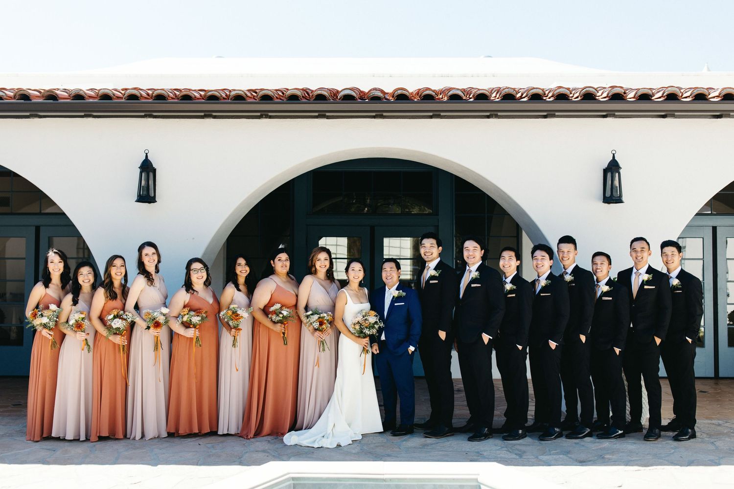 Wedding party poses together outside venue, bridesmaids in ombre rust and blush gowns with groomsmen in black suits.
