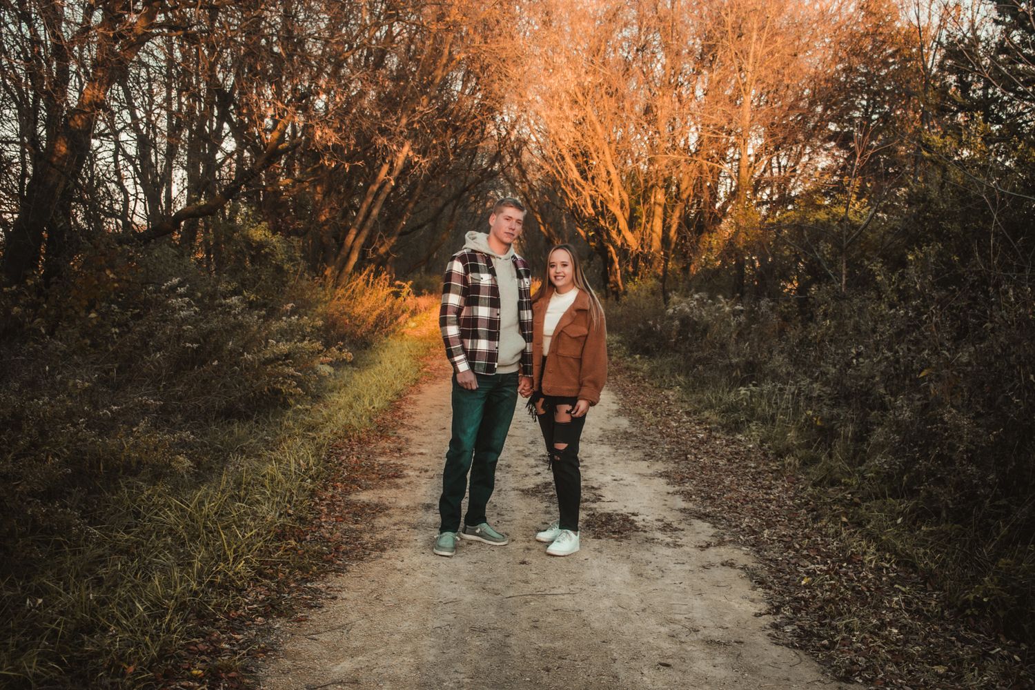 A couple stands together on a forest path with warm orange sunset lighting filtering through trees.