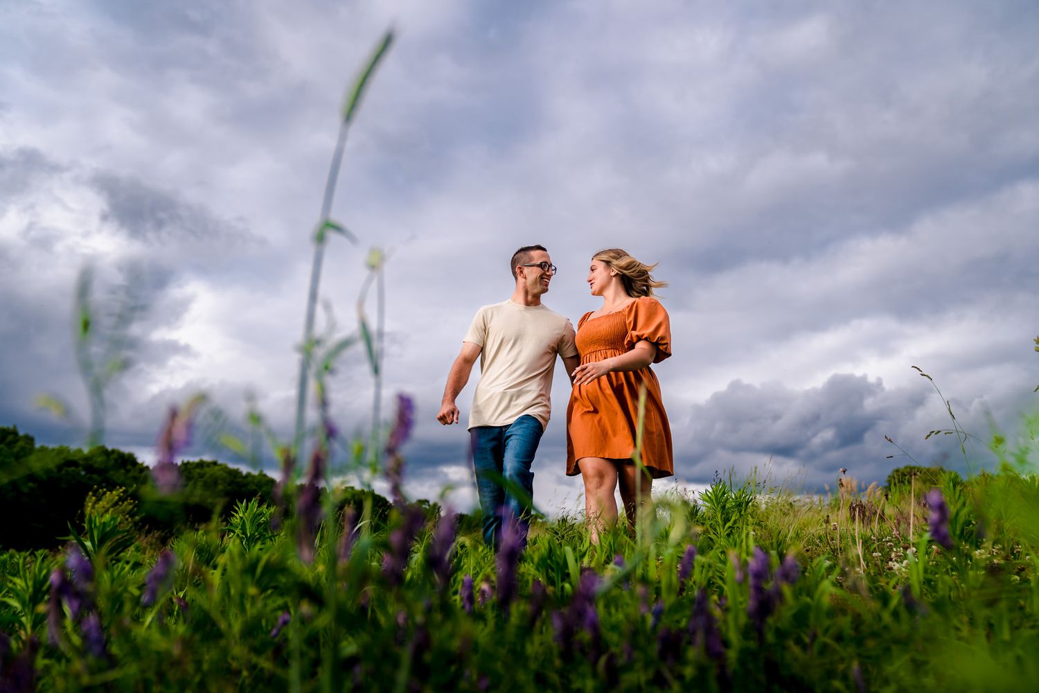 Two people stand together in a wildflower meadow with purple blooms under cloudy skies.