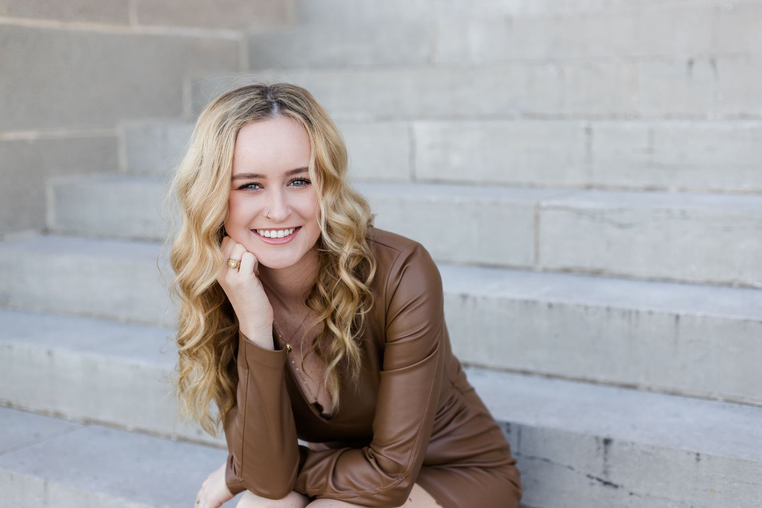 Woman in brown leather jacket smiles while sitting on concrete steps outdoors.