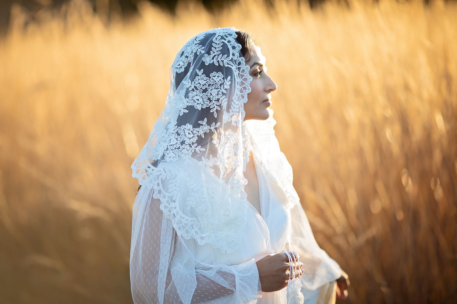 Woman in white lace chapel veil standing in golden wheat field in soft sunset light.