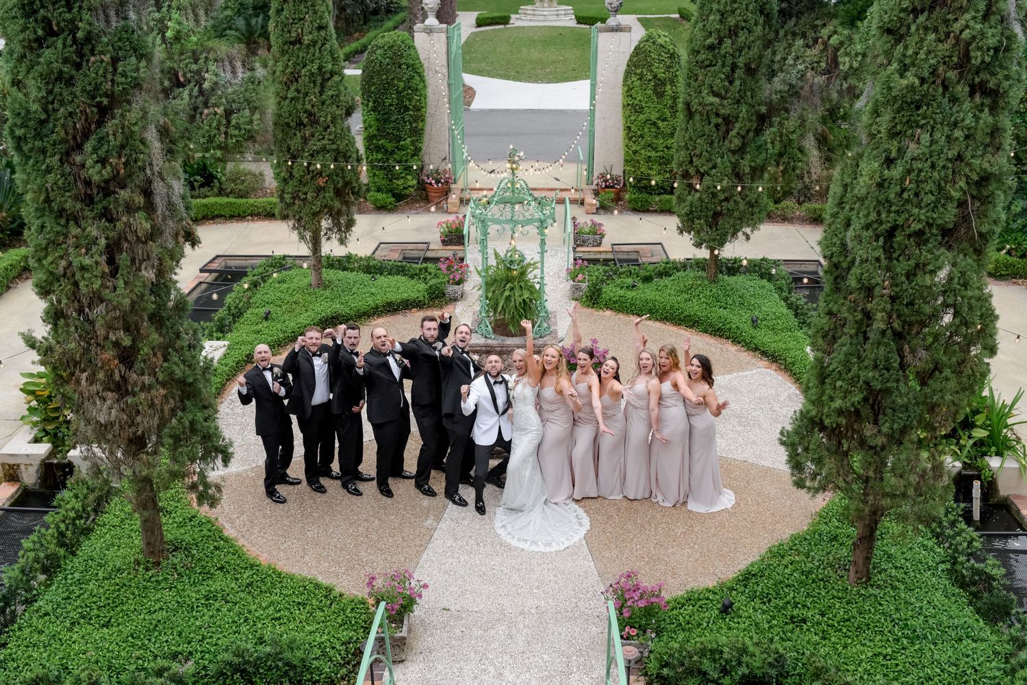Wedding party poses in formal attire around a garden gazebo surrounded by cypress trees.