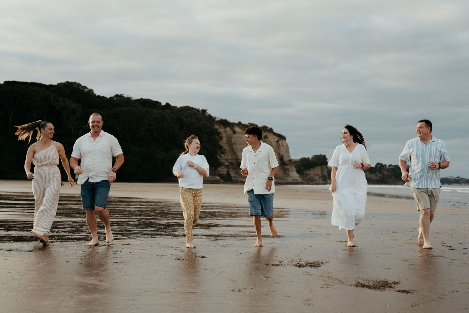 Family of six walking and laughing together on a sandy beach with rocky cliffs in the background.