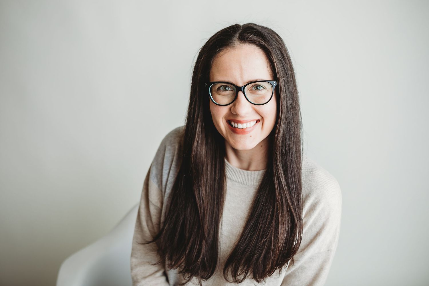 Portrait of someone with long dark hair and glasses wearing a beige sweater against grey wall.