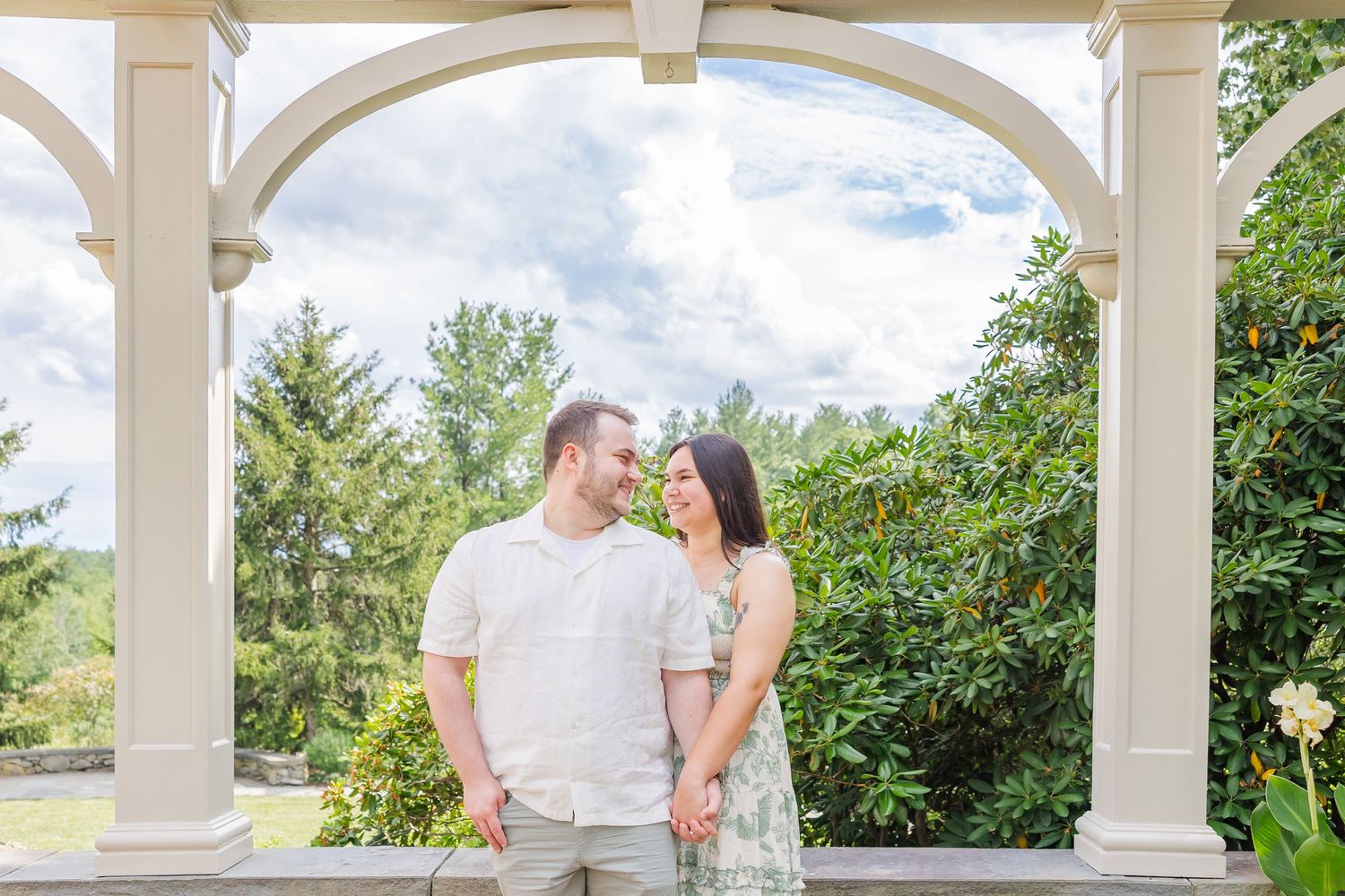 Couple embracing and smiling at each other in front of a lush green wall covered in colorful pink and yellow flowers.