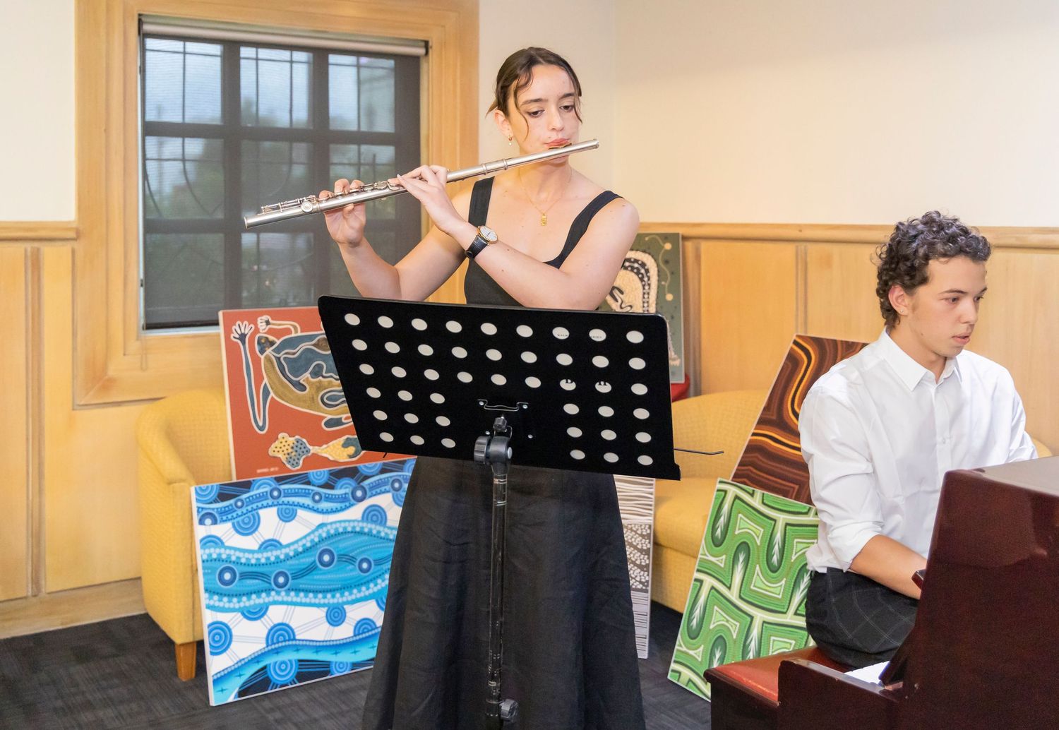 Musicians perform with flute and piano in a wood-paneled room with colorful artwork.