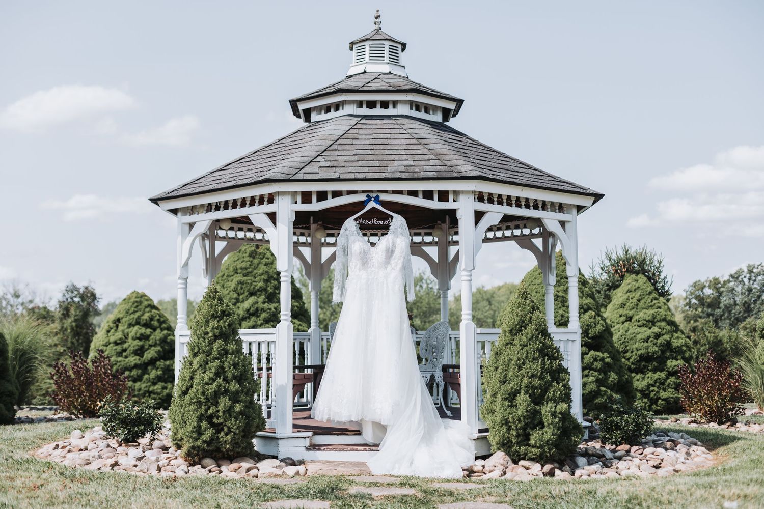 Elegant wedding dress hanging in a white gazebo surrounded by evergreen trees on a sunny day.