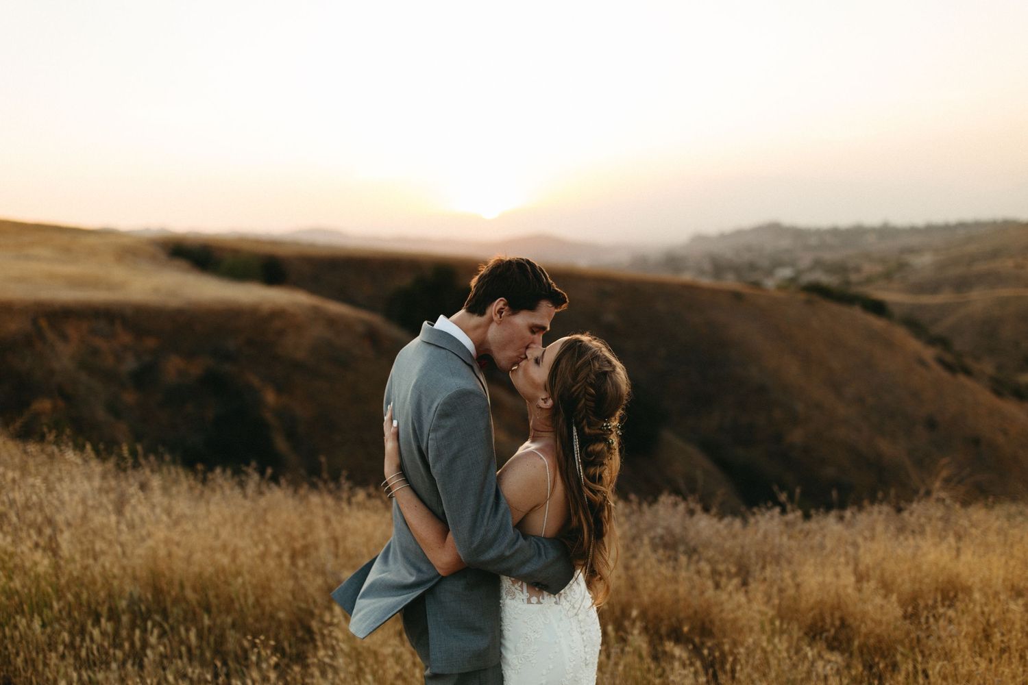 A couple shares a romantic kiss in a golden field of tall grass at sunset.