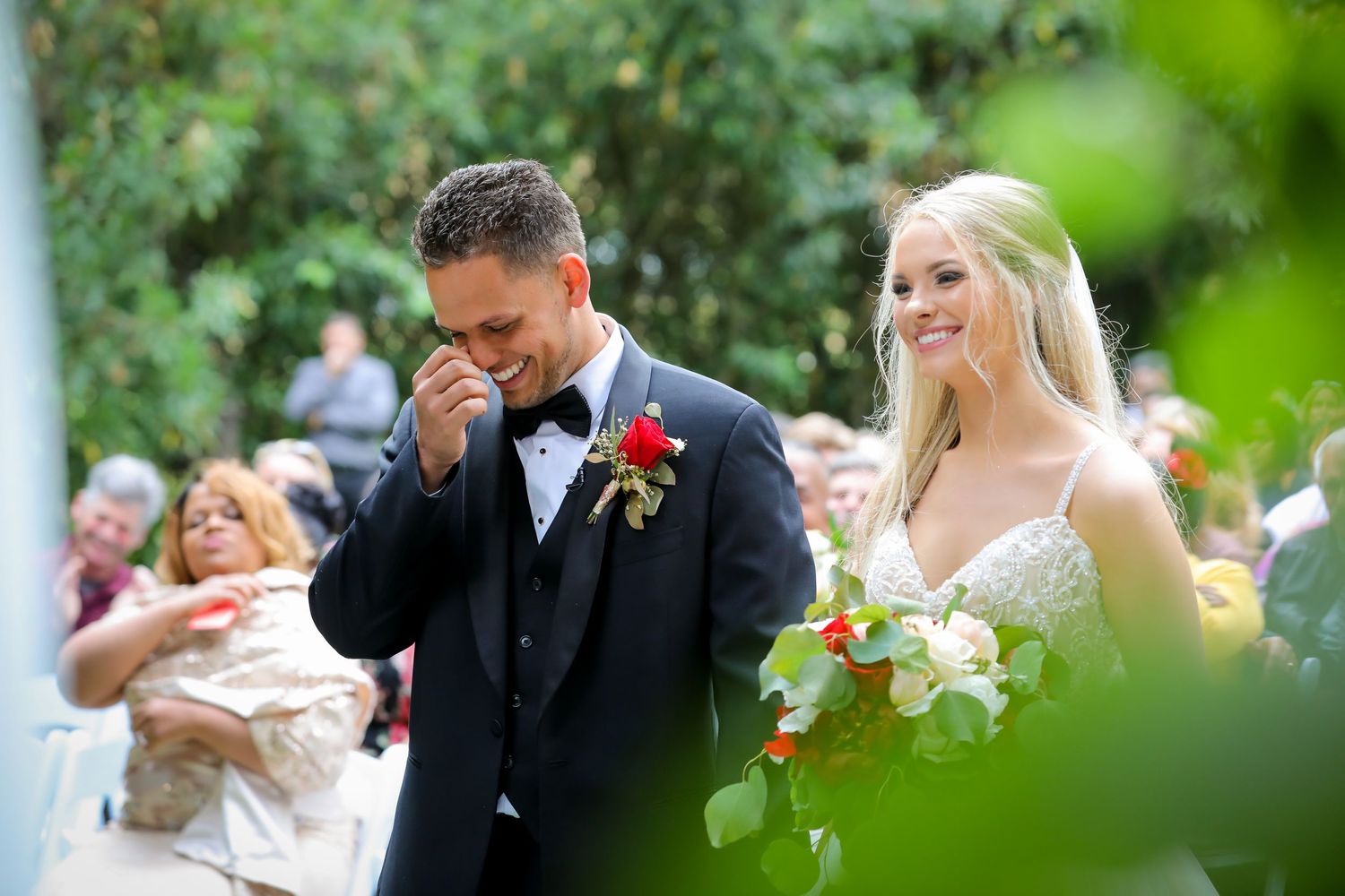 Emotional outdoor wedding ceremony moment with floral bouquet and greenery.