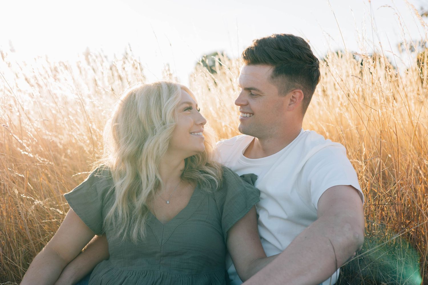 Couple smiles at each other while sitting in golden sunlit tall grass field.