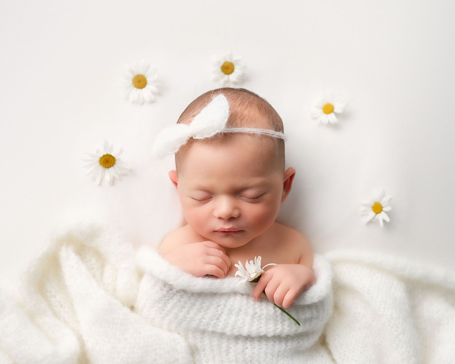 Serene newborn baby wrapped in white knit blanket holding daisy, surrounded by white flowers on soft background.